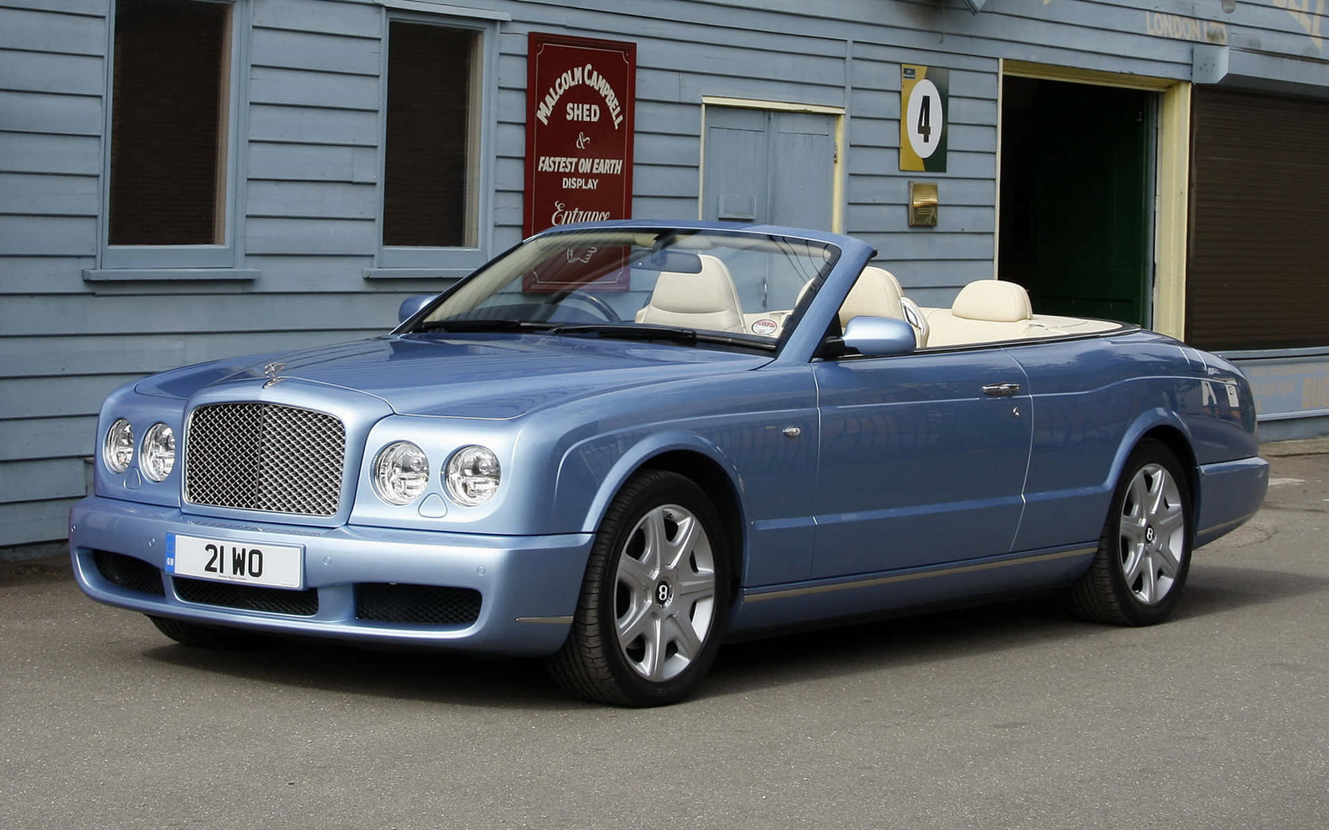 Bentley Azure 桌布 壁紙 — 一台時尚的淺藍色賓利敞篷車，搭配奶油色內裝，停在一個帶有復古賽車標誌的藍色木棚外。