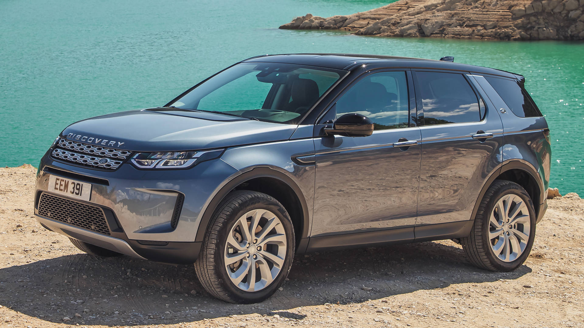 land rover iPhone 桌布 壁紙 — 一台時尚的灰色 Land Rover Discovery 停泊在陽光普照的岩石海岸旁，旁邊是碧綠色的海水。