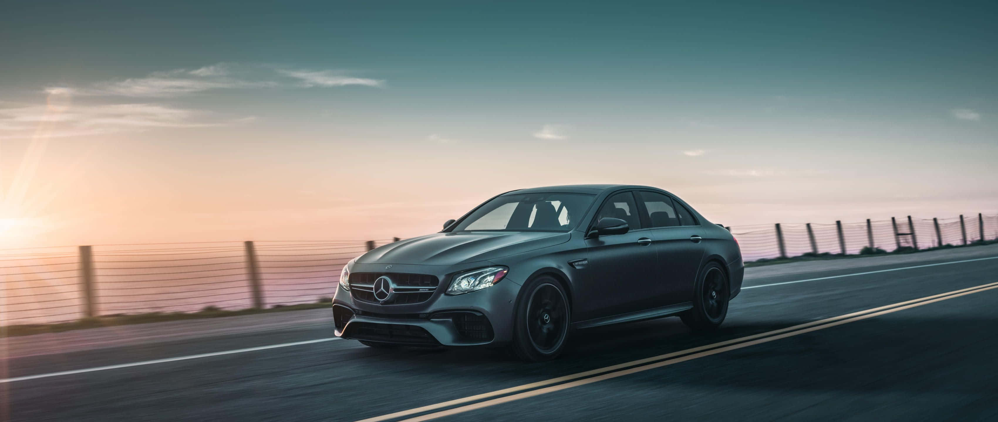 Mercedes 桌布 壁紙 — 一台時尚的灰色 Mercedes-AMG 轎車沿著海岸公路在日落時分疾馳，背後閃耀著溫暖的金色光芒。
