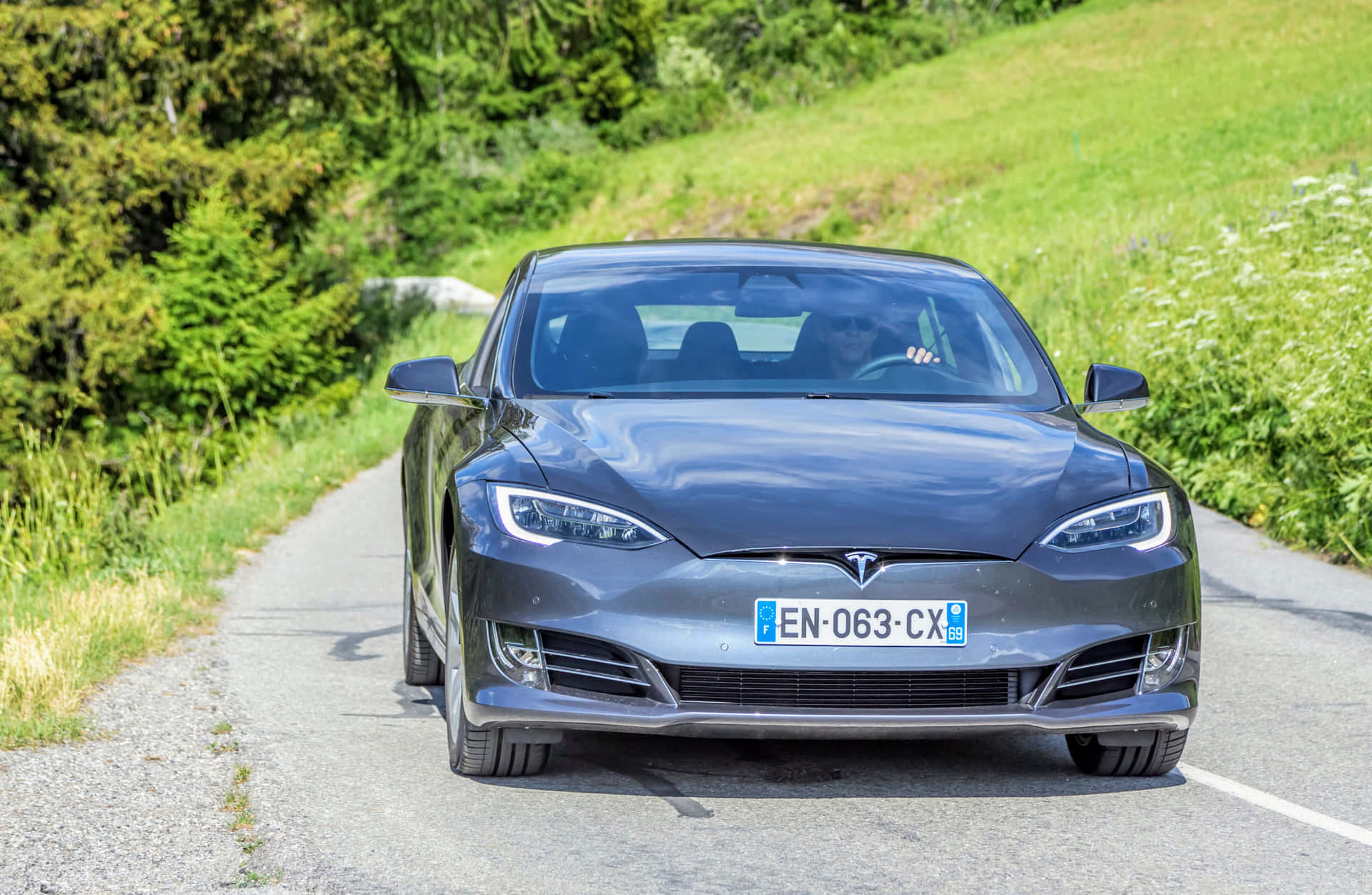 Tesla Car 壁紙 — 一台時尚的灰色 Tesla Model S 在蜿蜒的鄉村道路上向鏡頭駛來，兩旁是鬱鬱蔥蔥的綠色山丘和樹木。