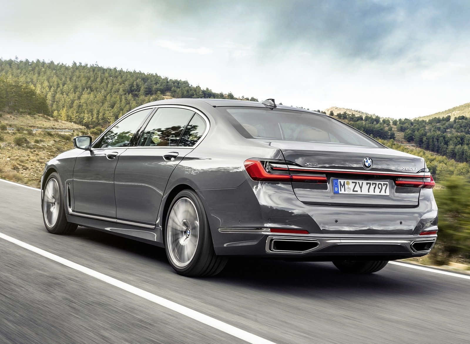 Bmw 7 Series 桌布 壁紙 — 一台時尚的灰色BMW 7系列轎車沿著蜿蜒的山路行駛，兩旁是森林覆蓋的山丘，天空多雲。