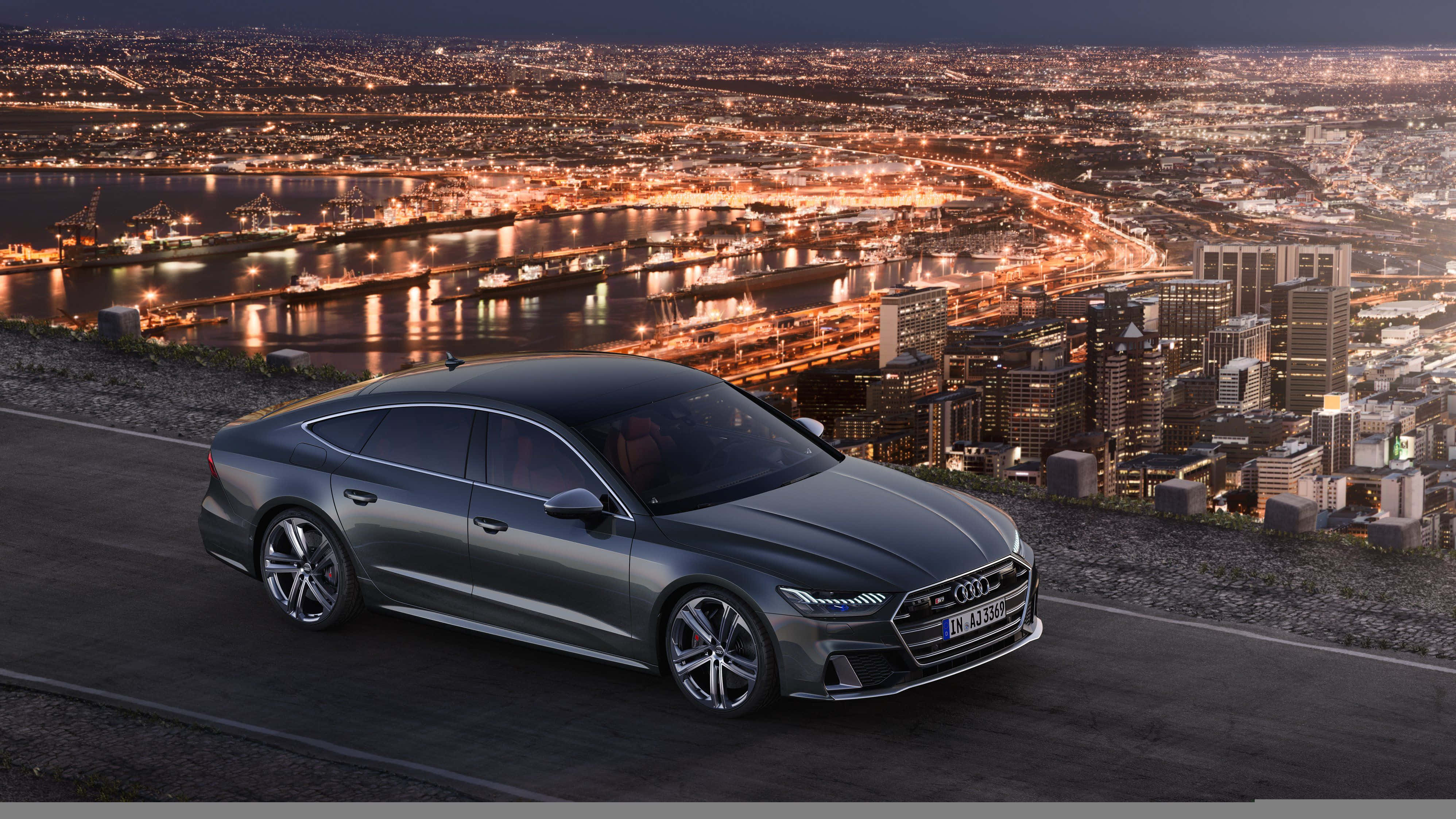 Audi S7 桌布 壁紙 — 一台時尚的灰色奧迪轎車停泊在山上，俯瞰夜晚充滿活力的城市天際線，閃爍的燈光倒映在水面上。