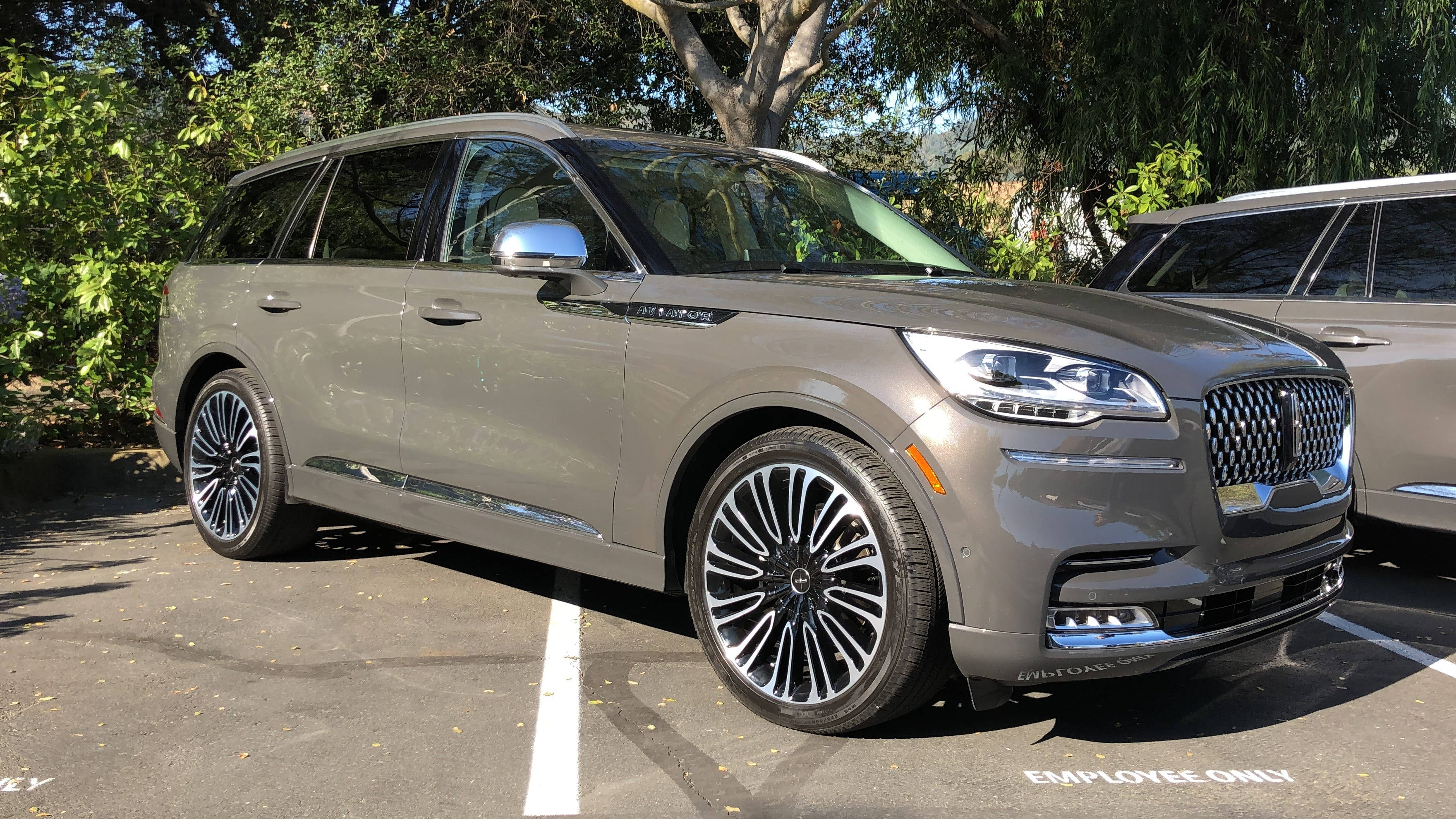 cars 桌布 壁紙 — 一台時尚的灰色林肯Navigator SUV停放在陽光充足的停車場，附近有樹木和另一輛車。