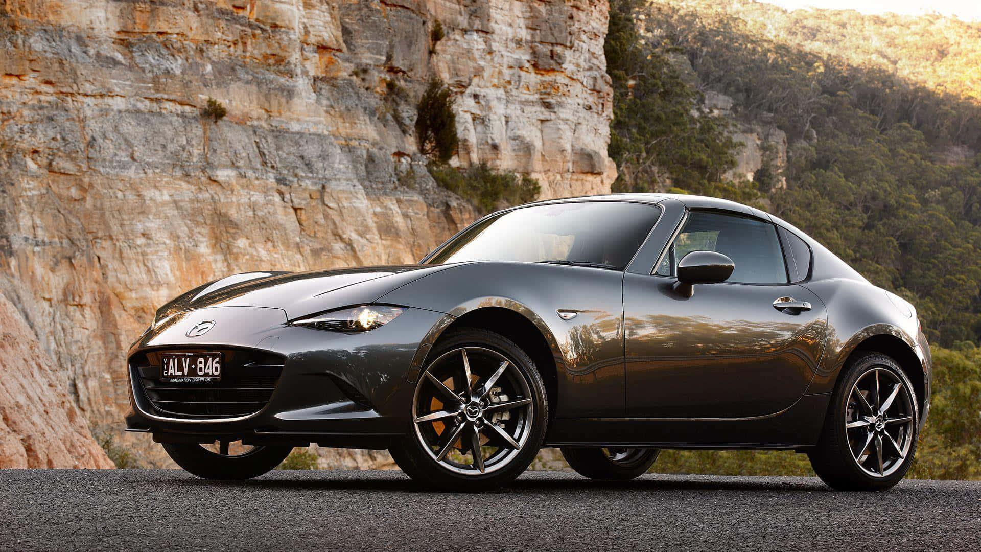 Miata 桌布 壁紙 — 一台時尚的灰色馬自達MX-5 Miata跑車停泊在崎嶇懸崖旁的柏油路上，金色陽光凸顯其曲線。