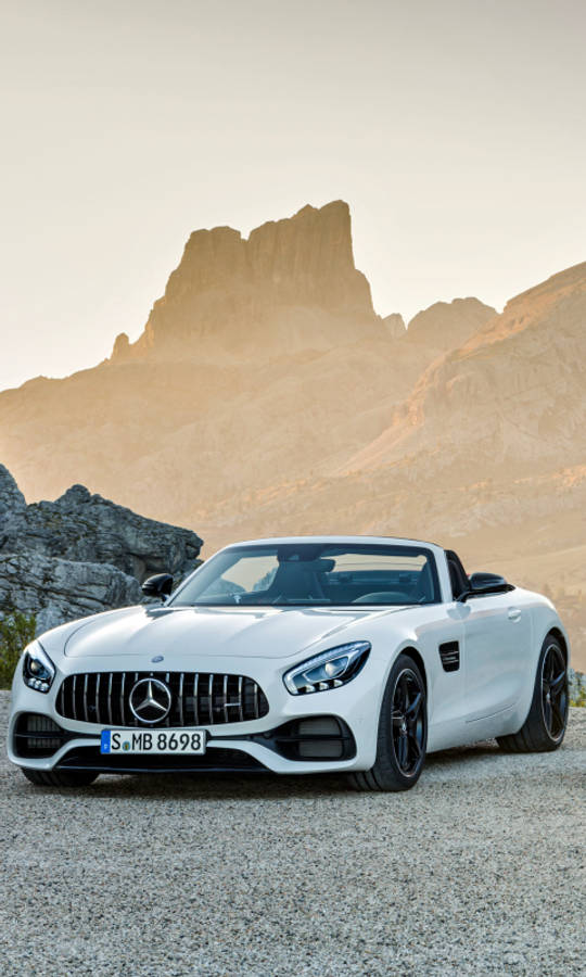 mercedes amg iPhone 桌布 壁紙 — 一台時尚的白色 Mercedes-AMG GT Roadster 停泊在碎石上，背景是沐浴在金色日落光芒中的戲劇性岩石山脈。