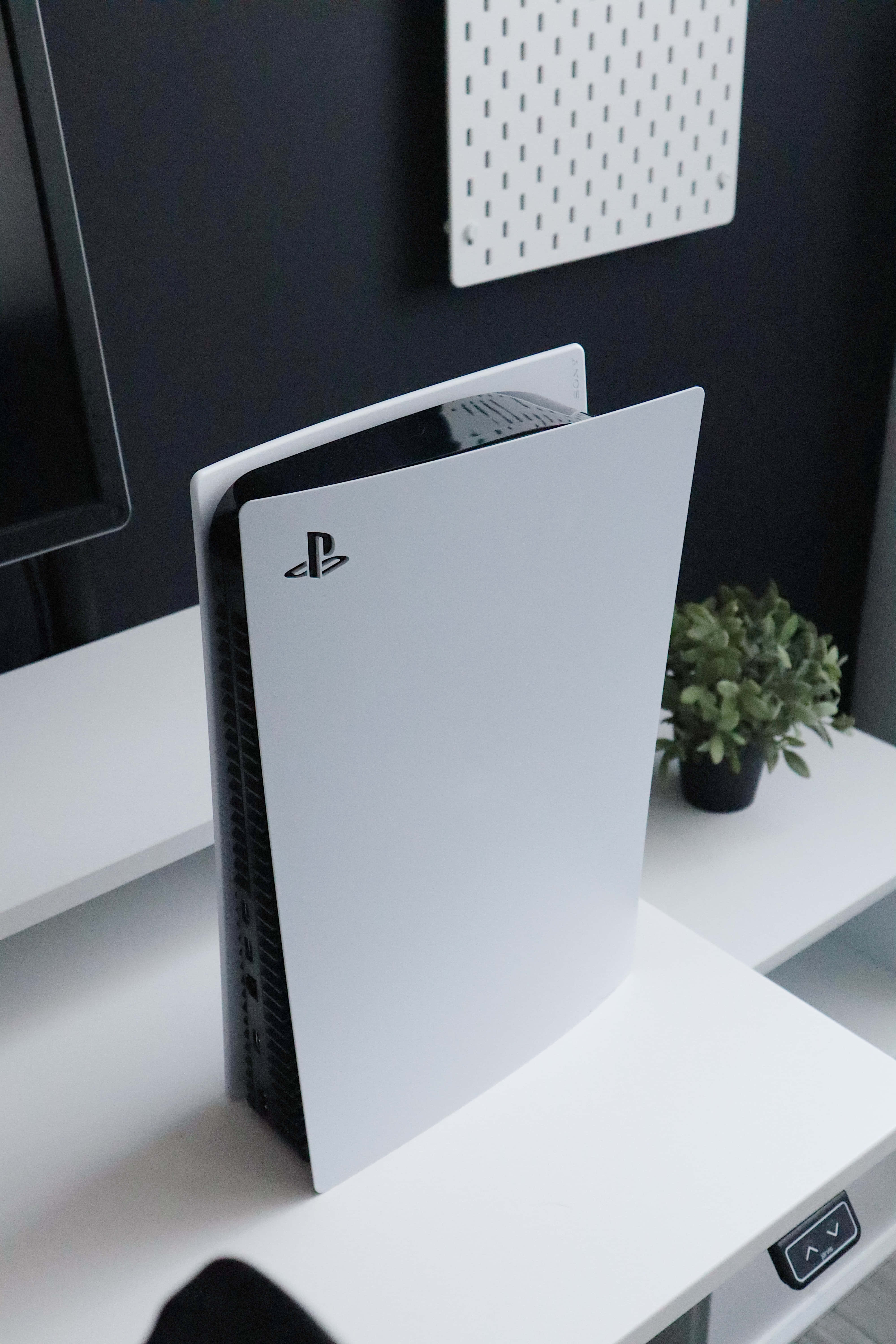 Gaming Consoles 桌布 壁紙 — 一台時尚的白色 PlayStation 5 主機直立放置在簡約的白色桌子上，旁邊有一棵小綠色植物和一個顯示器。
