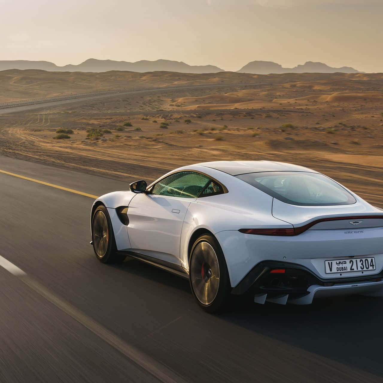 奧斯頓馬丁 桌布 壁紙 — 一台時尚的白色Aston Martin在沙漠高速公路上奔馳，金色沙丘和遠山映襯著溫暖的日落天空。