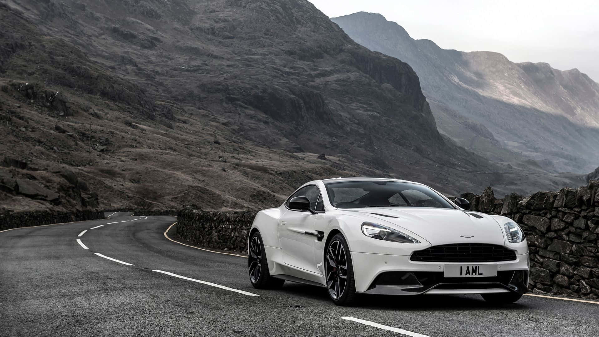 Aston Martin Vanquish 桌布 壁紙 — 一台時尚的白色Aston Martin跑車在蜿蜒的山路上行駛，兩旁是崎嶇的石牆和霧氣籠罩的山峰。