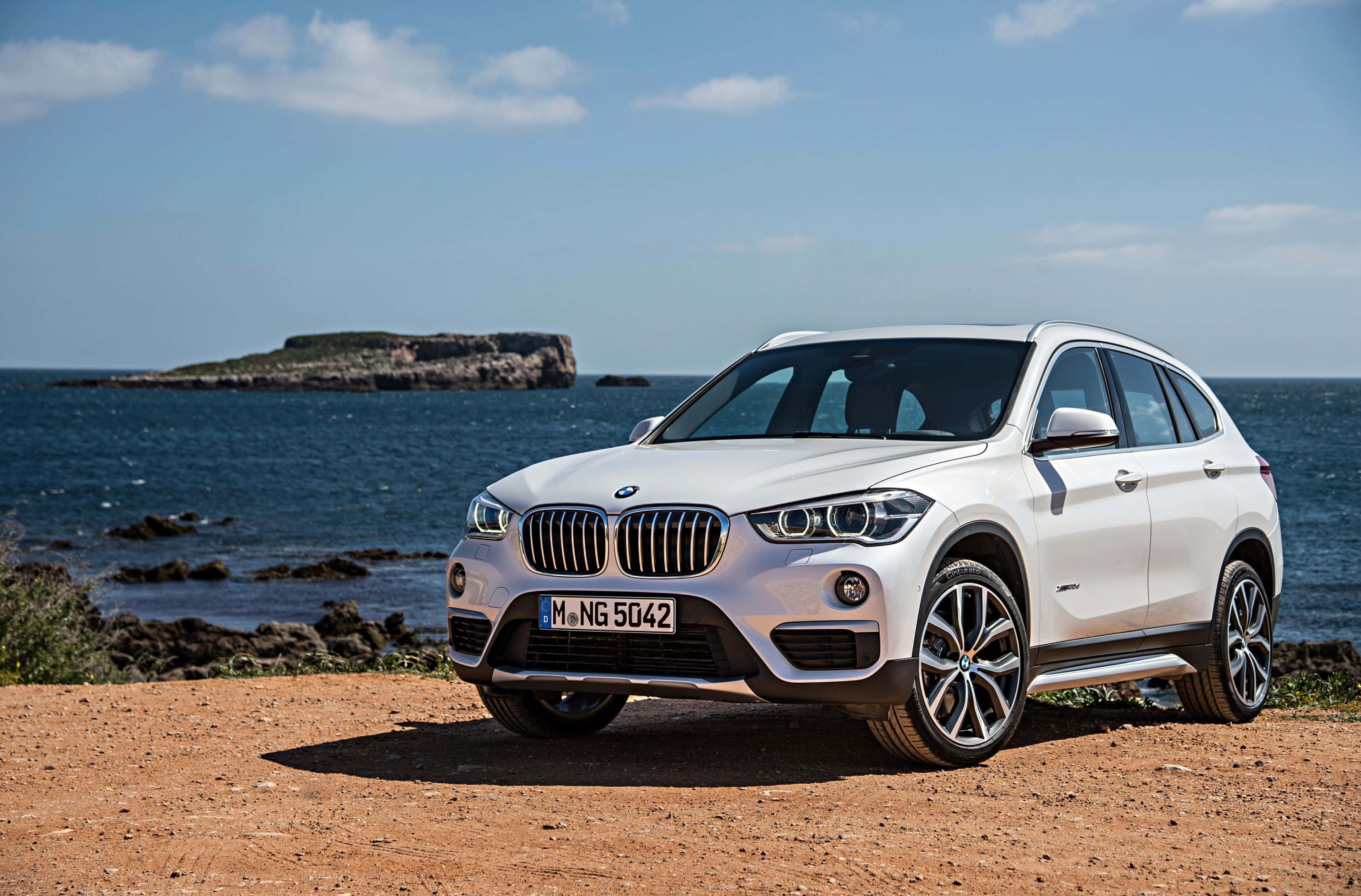 Bmw X1 桌布 壁紙 — 一台時尚的白色BMW X1 SUV停放在陽光明媚的海岸懸崖上，俯瞰深藍色的海洋，遠處有岩石小島。