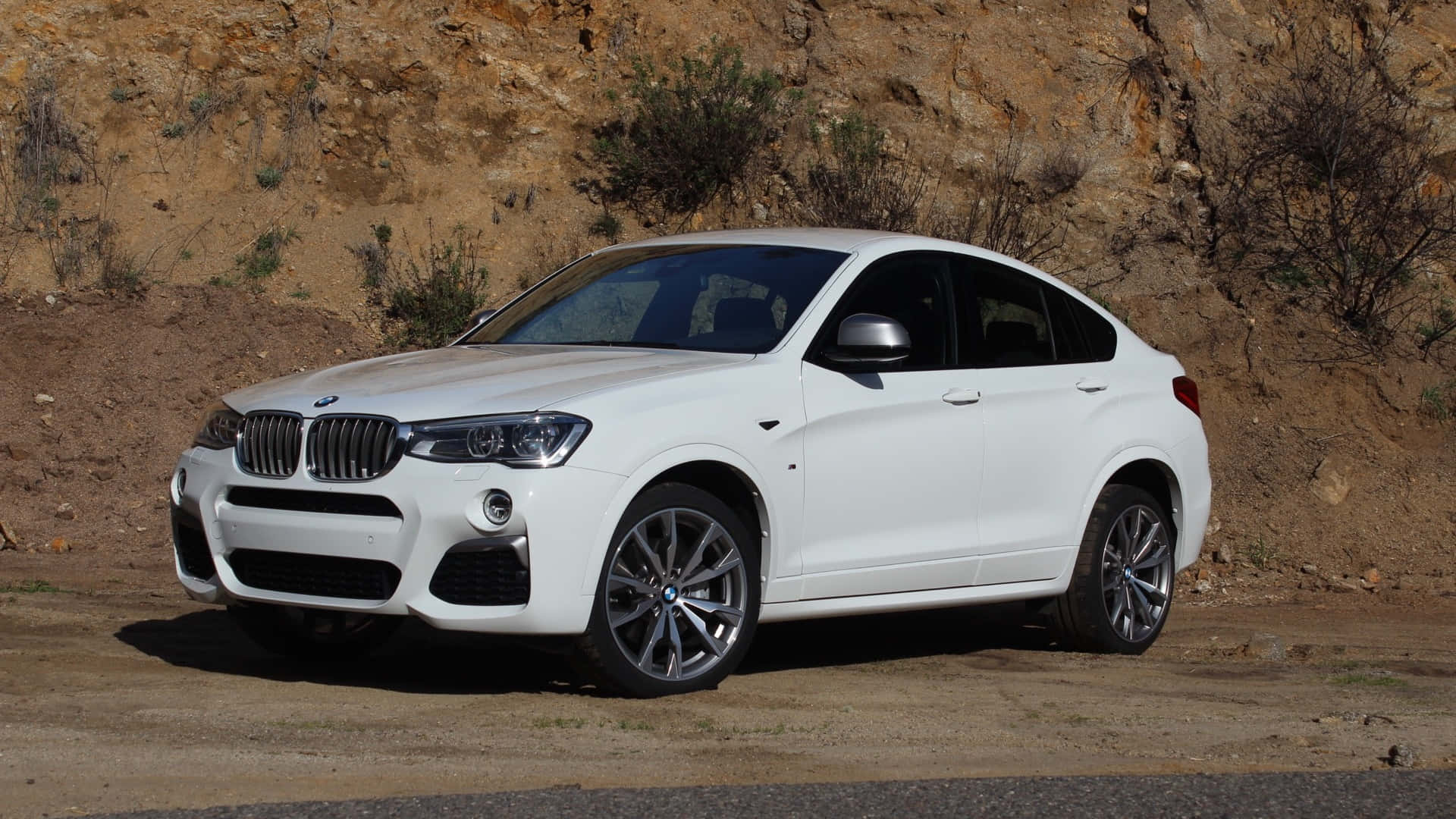 Bmw X4 桌布 壁紙 — 一台時尚的白色BMW X4轎跑SUV停泊在塵土飛揚的路邊，背景是崎嶇的棕色懸崖和稀疏的綠色灌木。