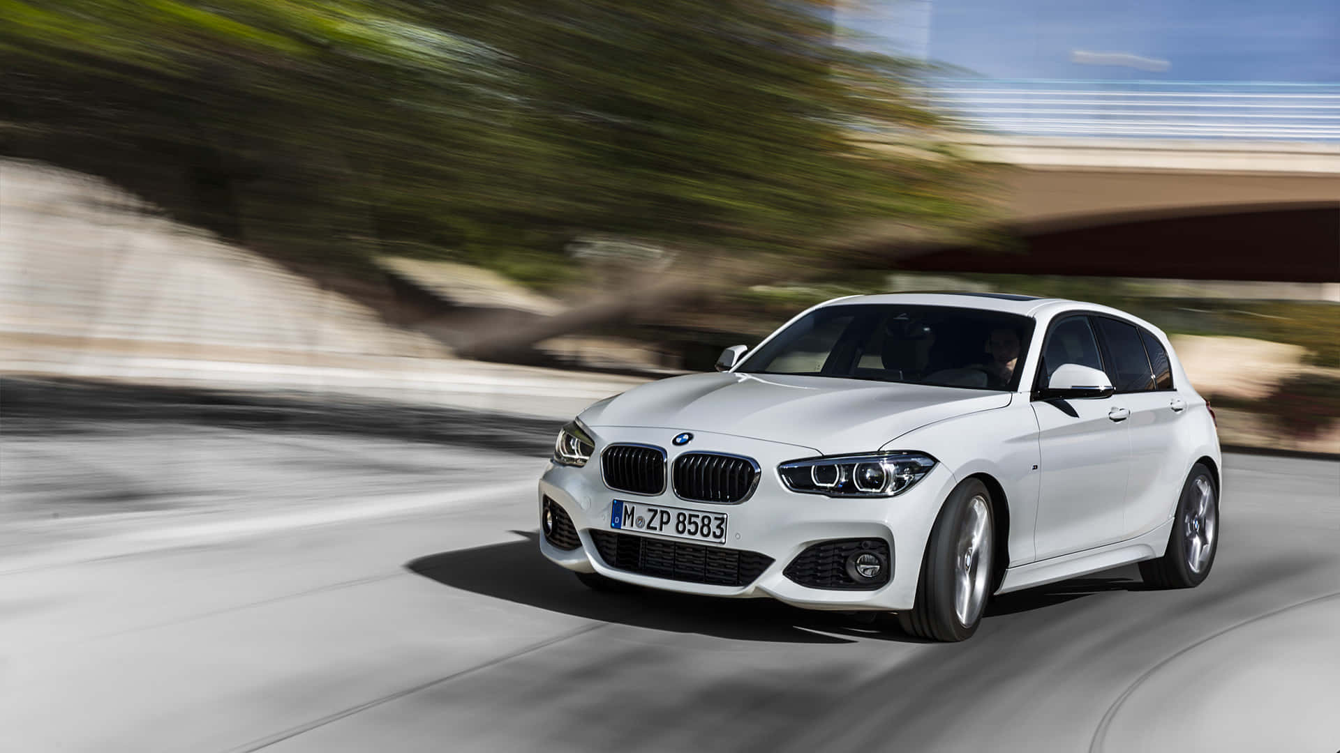 Bmw 1 Series 桌布 壁紙 — 一台時尚的白色BMW掀背車沿著彎曲的道路疾馳，上方有模糊的綠化和混凝土橋樑。