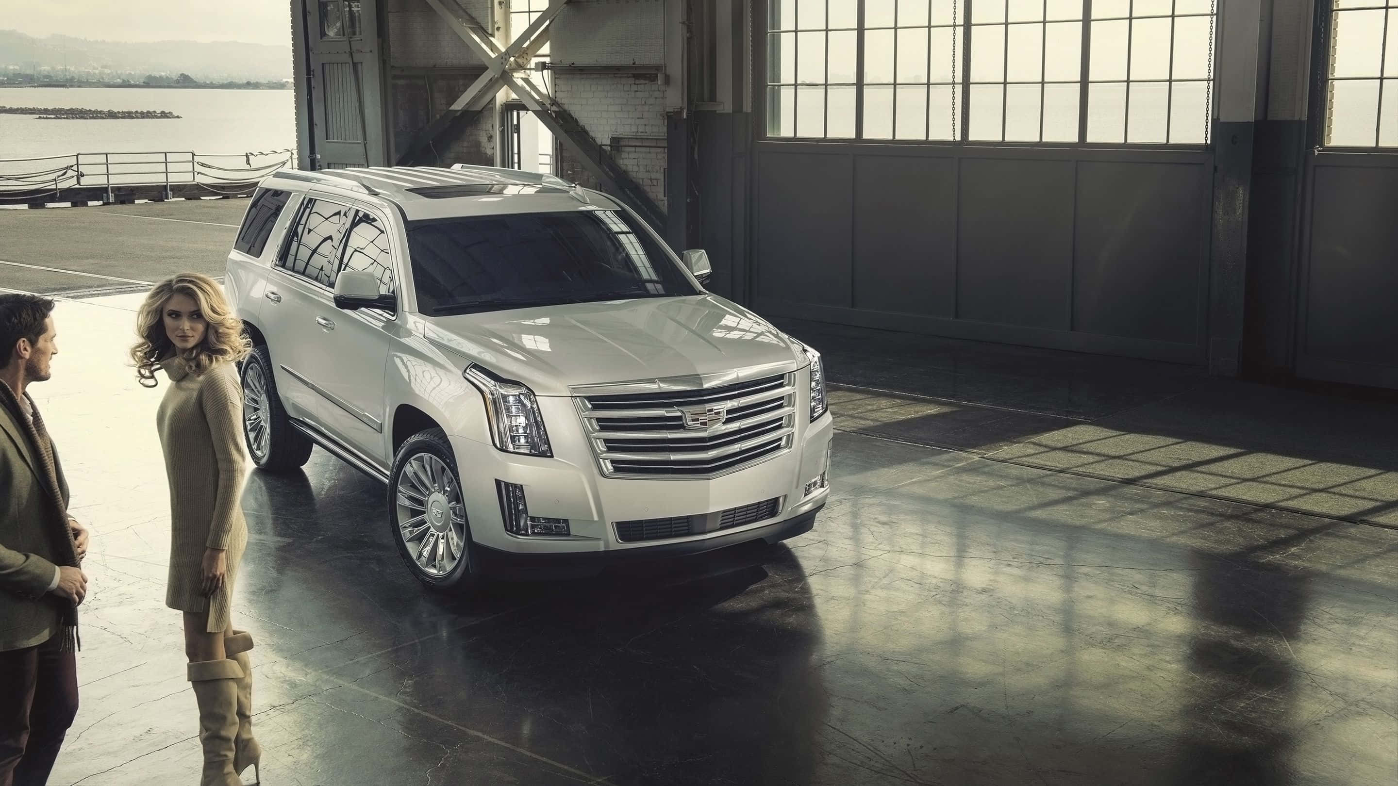 Cadillac Escalade 桌布 壁紙 — 一台時尚的白色凱迪拉克SUV在自然光下，於寬敞的工業機庫中閃閃發光，旁邊站著兩位衣著優雅的人。