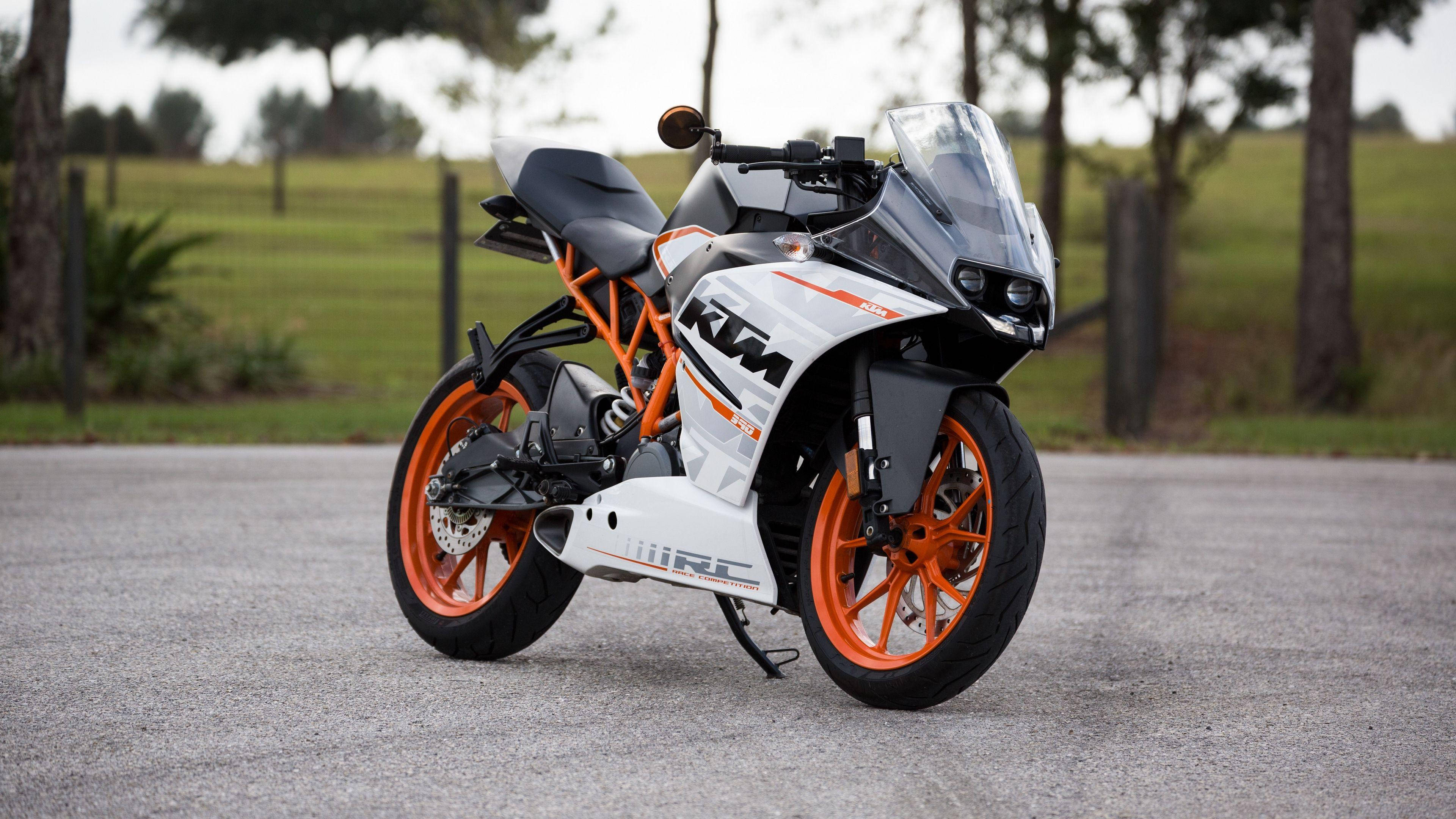 機車 桌布 壁紙 — 一台時尚的白色和橙色KTM RC機車停在碎石路上，周圍是綠草和樹木。