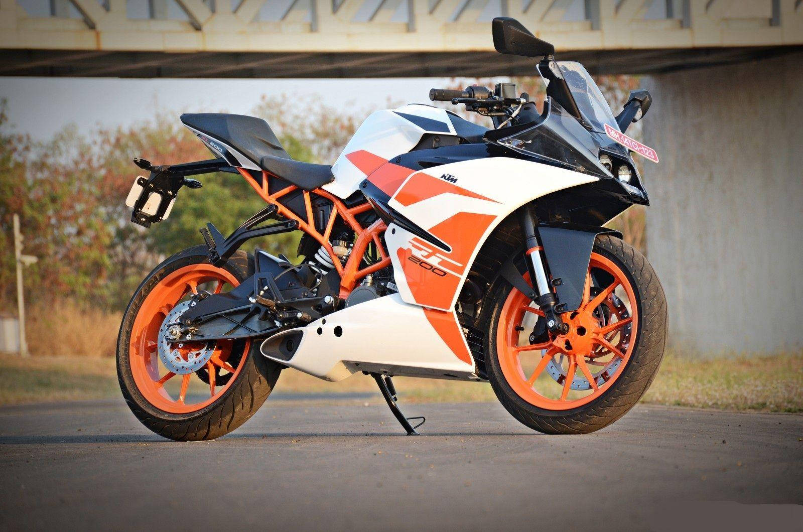 KTM RC 200 桌布 壁紙 — 一台時尚的白色和橙色KTM運動機車停放在混凝土橋下，背景有秋天的樹木。