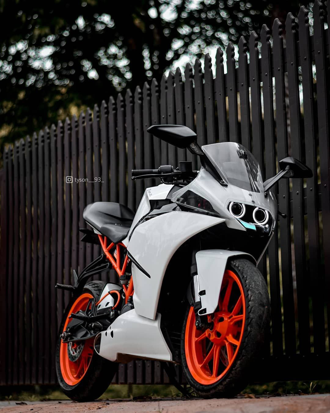 KTM RC 200 桌布 壁紙 — 一台時尚的白色和橙色運動機車，帶有黑色點綴，停在深色木柵欄旁，光線柔和。