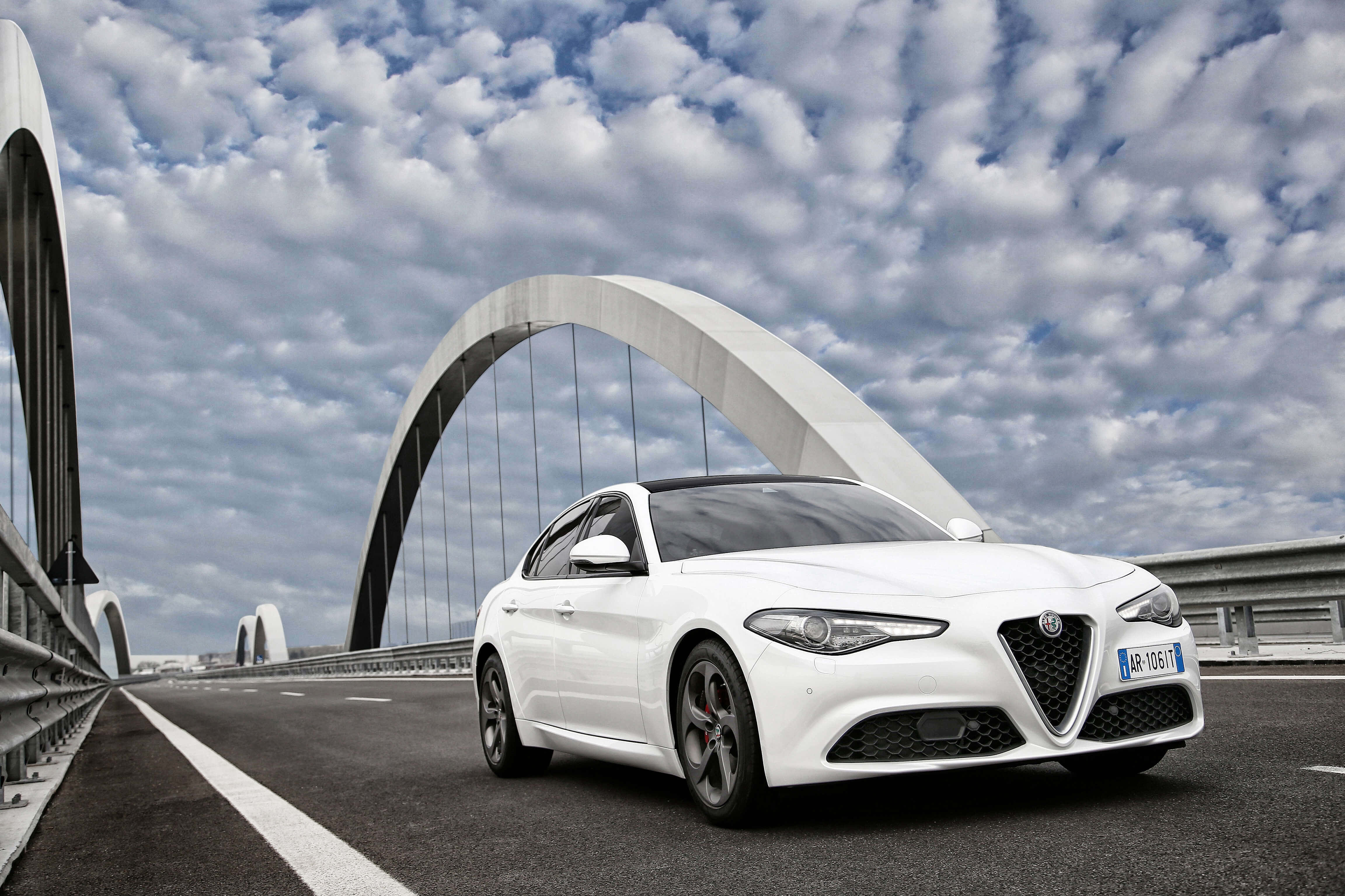 Alfa Romeo Giulia 桌布 壁紙 — 一台時尚的白色愛快羅密歐轎車在戲劇性的多雲天空下自信地行駛在現代拱橋上。