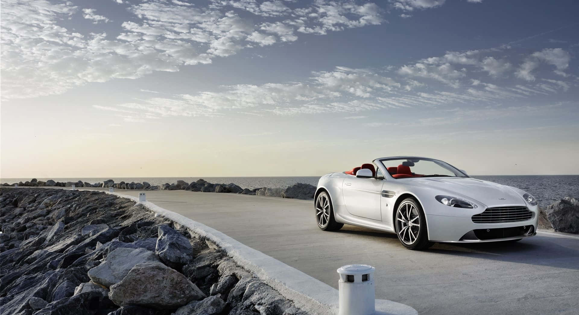 Aston Martin V12 Vantage 桌布 壁紙 — 一台時尚的白色敞篷跑車，配備紅色內裝，沿著海岸道路行駛，旁邊是岩石峭壁和海洋，天空部分多雲。