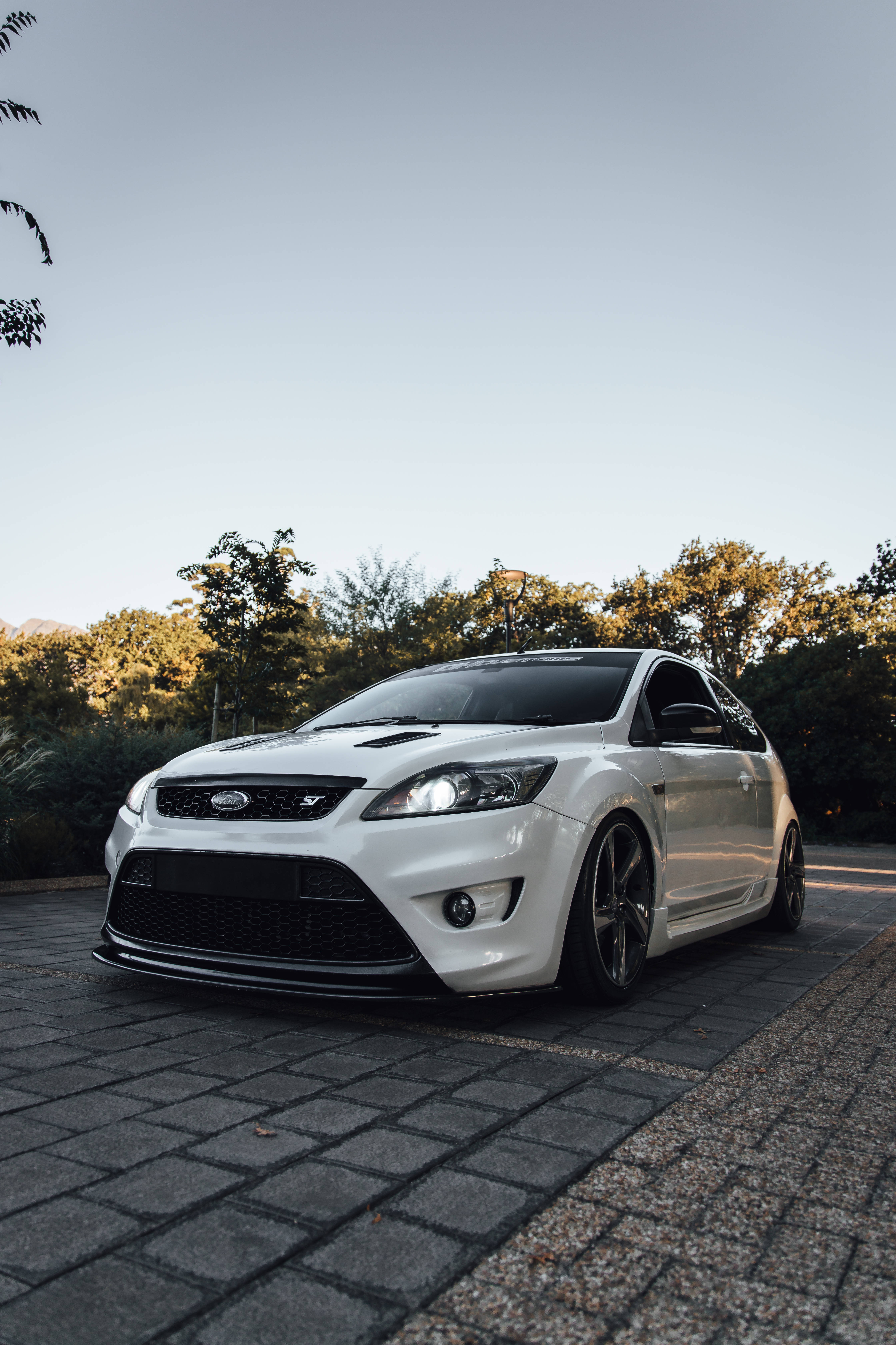 ford iPhone 桌布 壁紙 — 一台時尚的白色福特Focus ST停泊在鵝卵石小徑上，背景有樹木和柔和的金色光線。