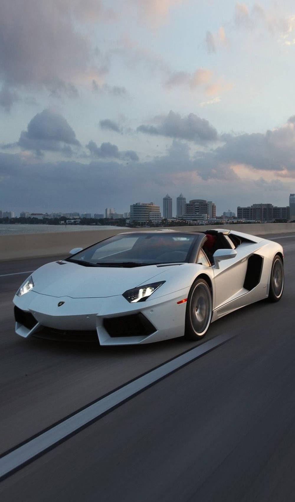 Amazing 桌布 壁紙 — 一台時尚的白色藍寶堅尼 Aventador 敞篷跑車在黃昏時分沿著海岸高速公路奔馳，遠處是城市天際線。