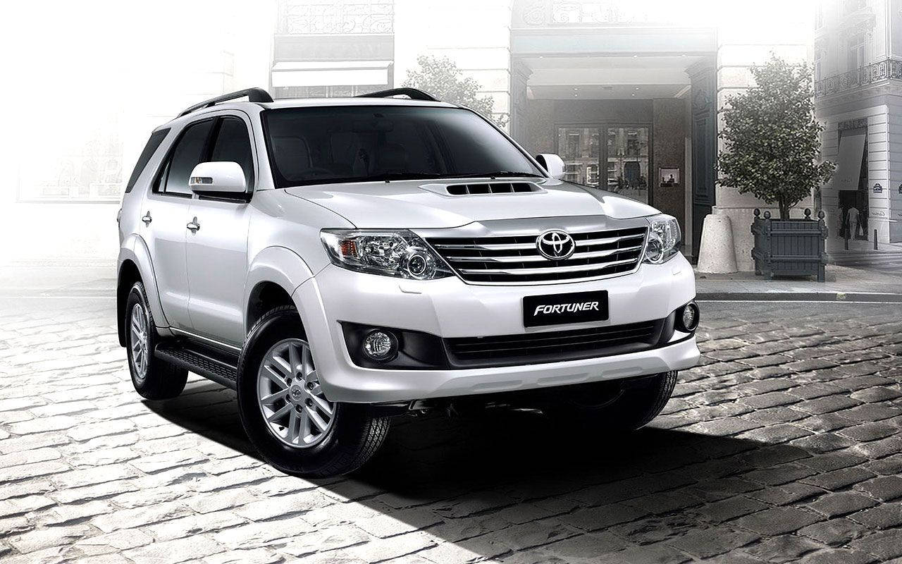 Toyota 桌布 壁紙 — 一台時尚的白色豐田 Fortuner SUV 停泊在鵝卵石街道上，前方是經典建築，沐浴在柔和的晨光中。