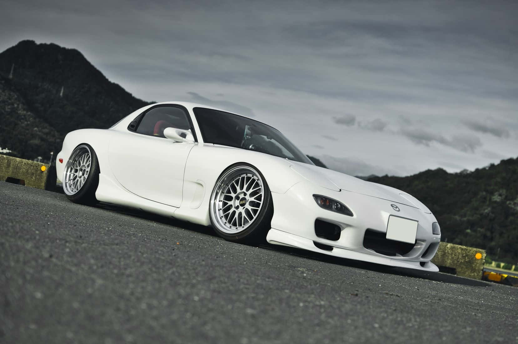 Mazda 桌布 壁紙 — 一台時尚的白色馬自達RX-7跑車配備定制輪圈，低趴在山路上，天空多雲。