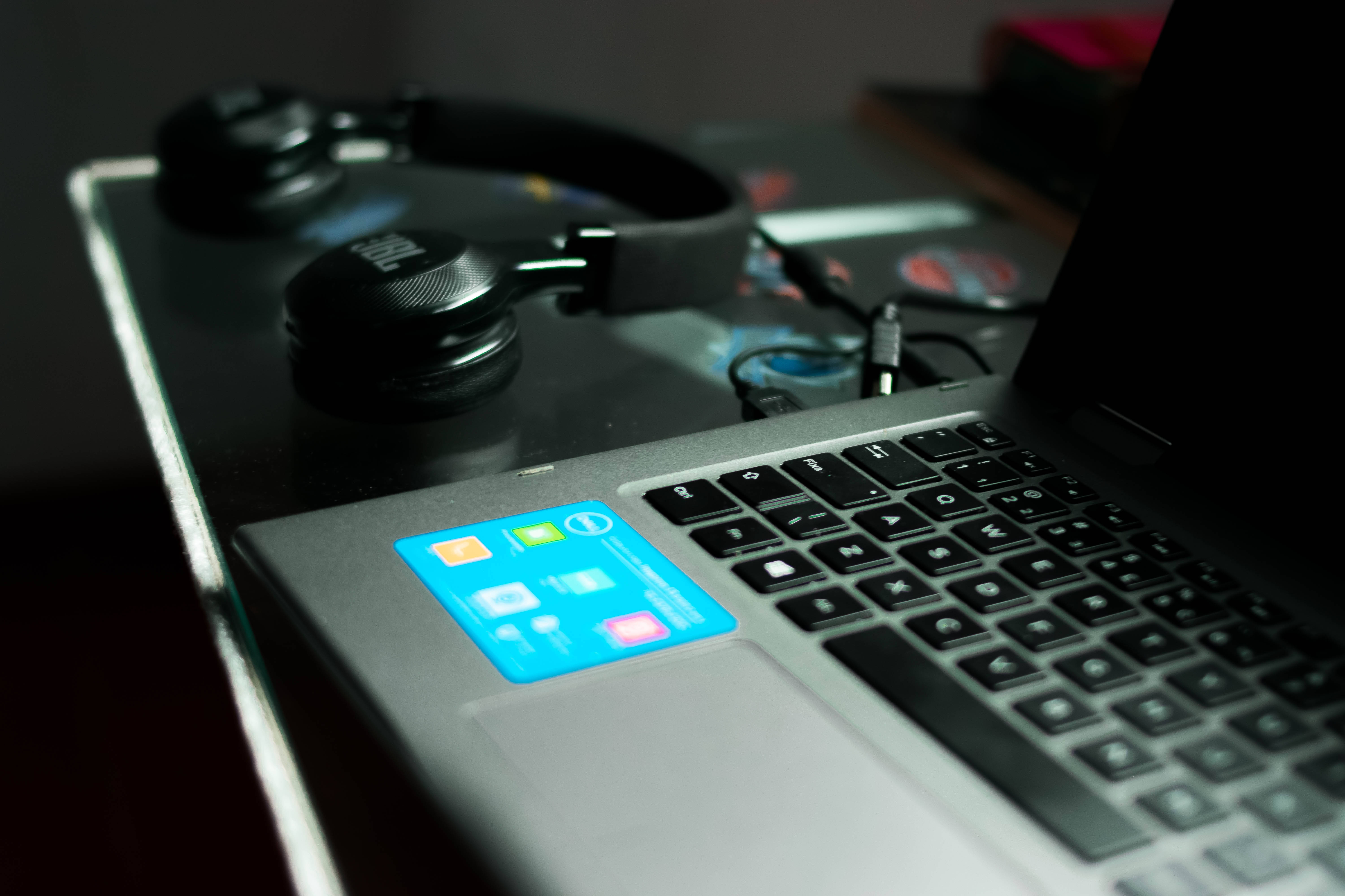 aesthetic tumblr laptop 桌布 壁紙 — 一台時尚的筆記型電腦，帶有發光的藍色應用程式圖標，旁邊放著黑色耳機，置於昏暗光線下的深色玻璃桌上。