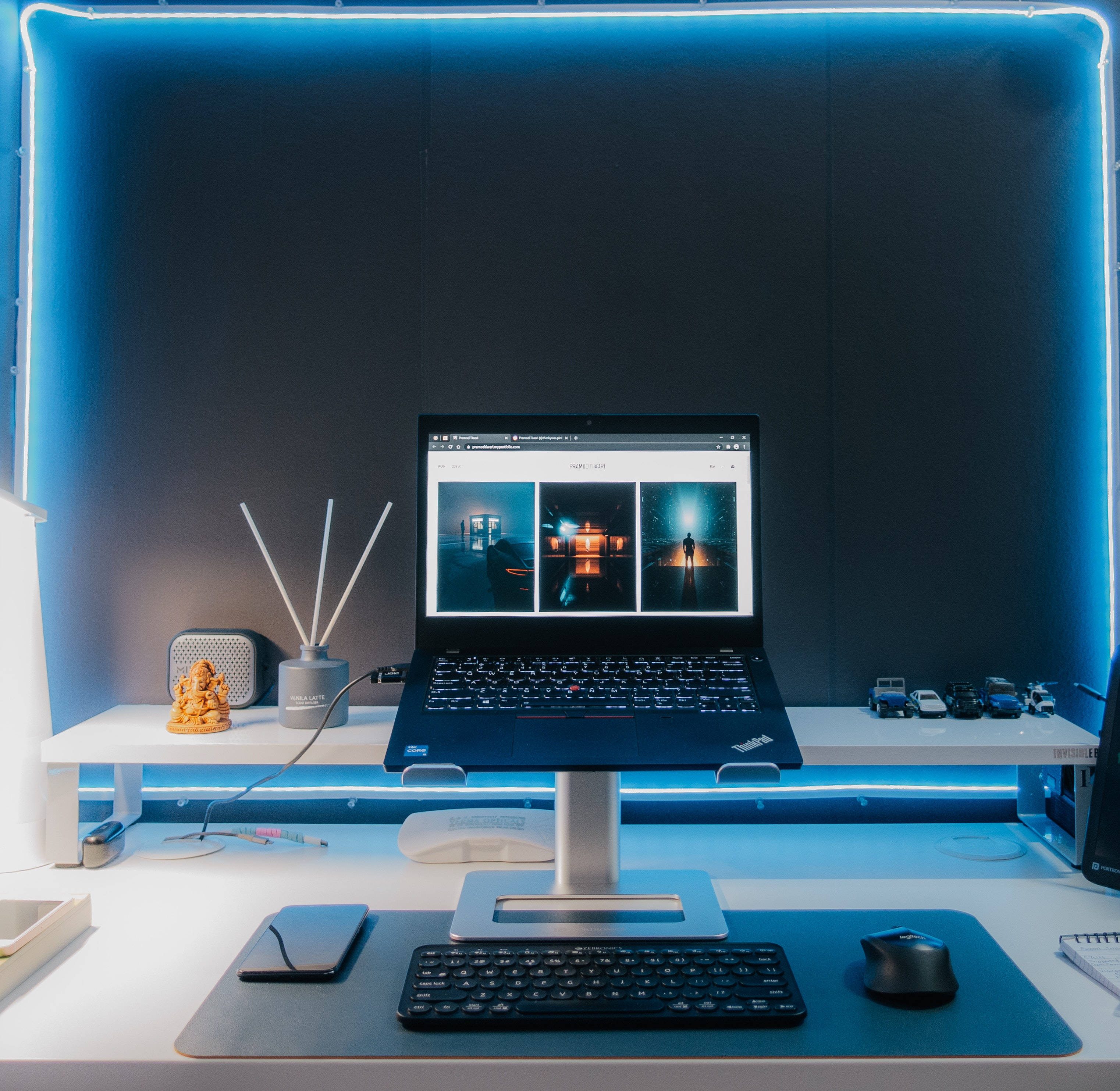 aesthetic tumblr laptop 桌布 壁紙 — 一台時尚的筆記型電腦放在支架上，顯示三個充滿氛圍的夜景，而發光的藍色霓虹燈框出一個簡約的白色桌面設置。