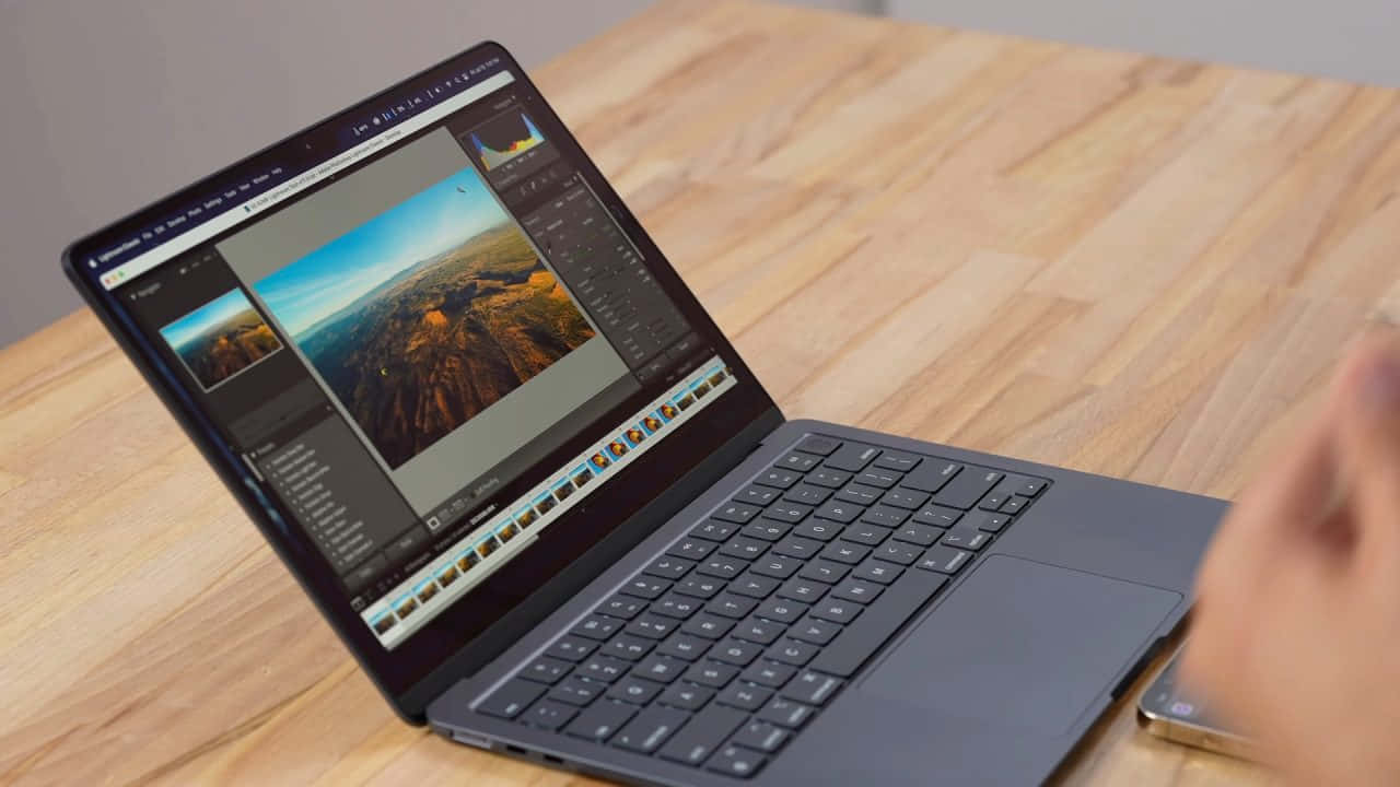 Black Macbook 桌布 壁紙 — 一台時尚的筆記型電腦放在木桌上，顯示一張正在軟體中編輯的鮮豔山景照片，旁邊有一隻手。