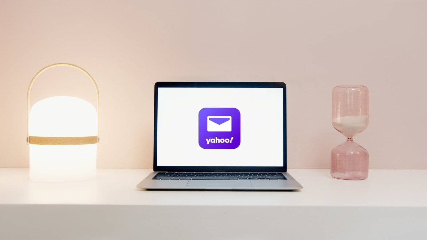 Yahoo Mail 桌布 壁紙 — 一台時尚的筆記型電腦顯示Yahoo標誌，置於白色書桌中央，兩側有發光的燈和粉紅色沙漏，背景是柔和的粉紅色牆壁。