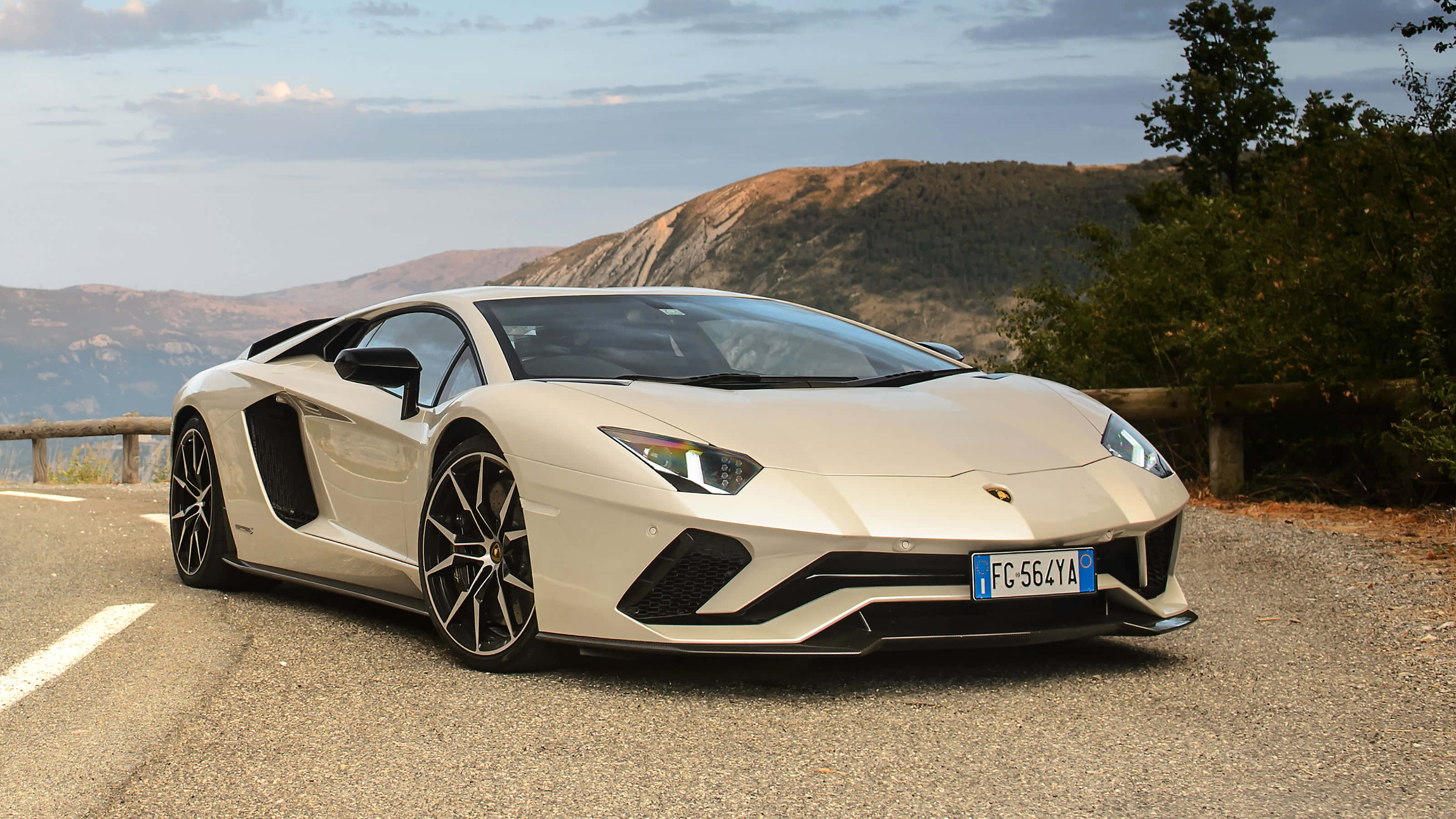 Cool Lamborghinis 桌布 壁紙 — 一台時尚的米色藍寶堅尼Aventador停泊在山路上，背景是崎嶇的山丘和樹木，天空多雲。