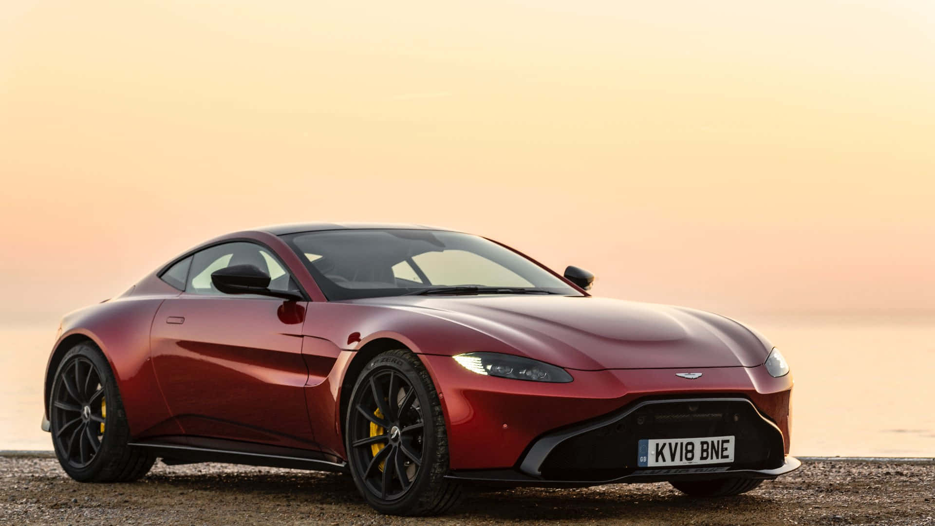 Aston Martin Vantage 桌布 壁紙 — 一台時尚的紅色Aston Martin跑車在日落時分的鵝卵石海灘上閃耀著溫暖的金色光芒。