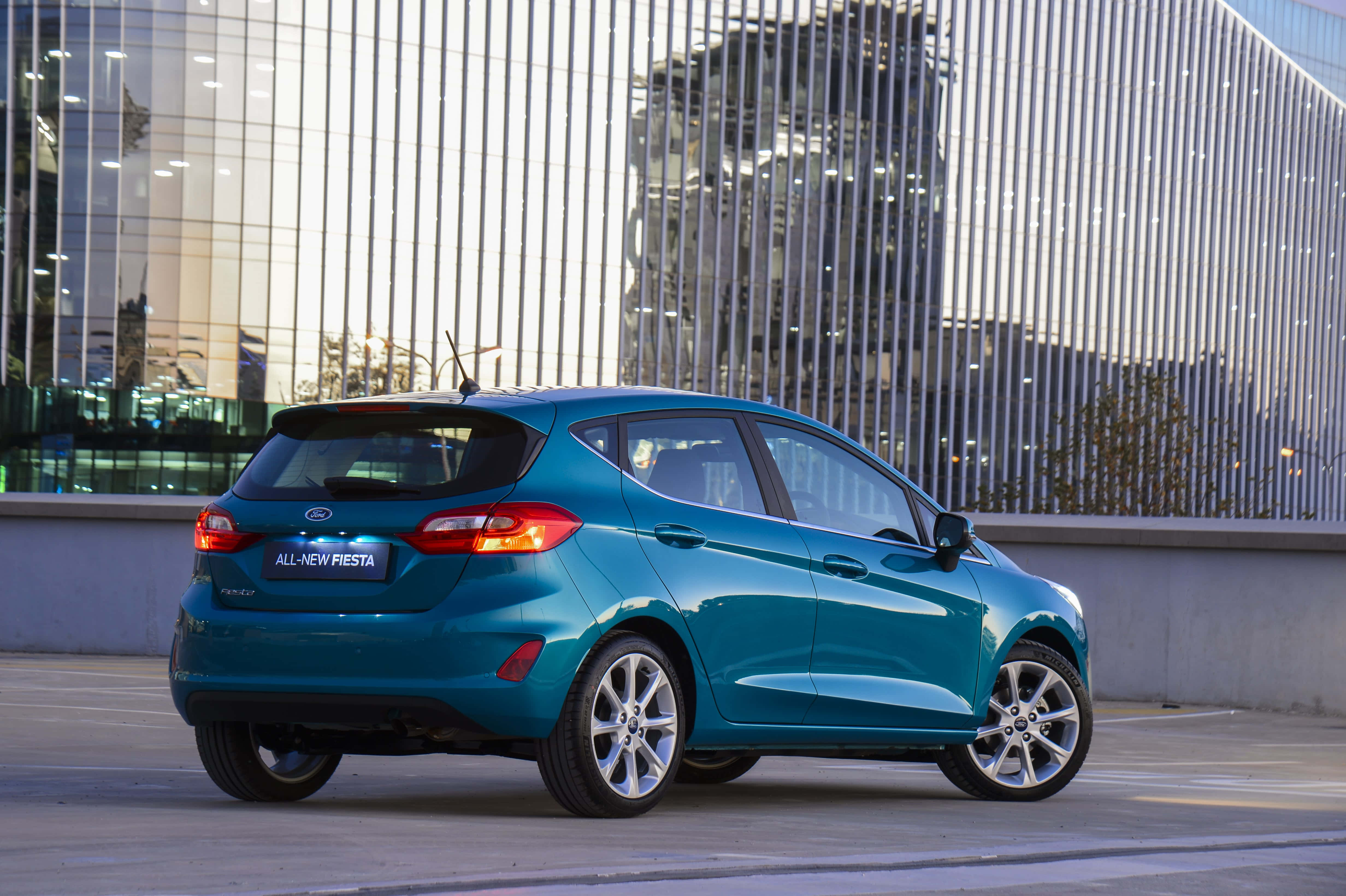 Compact Car 桌布 壁紙 — 一台時尚的藍綠色福特 Fiesta 停放在現代玻璃建築前，建築帶有垂直金屬框架。