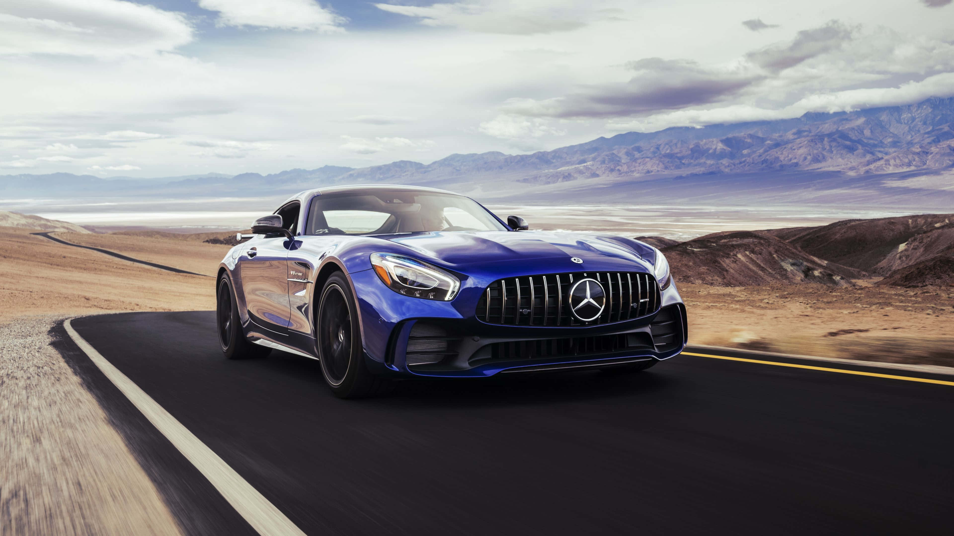 4K 奔馳壁紙 桌布 壁紙 — 一台時尚的藍色 Mercedes-AMG GT 跑車在沙漠公路上疾馳，遠方山巒起伏，天空多雲。
