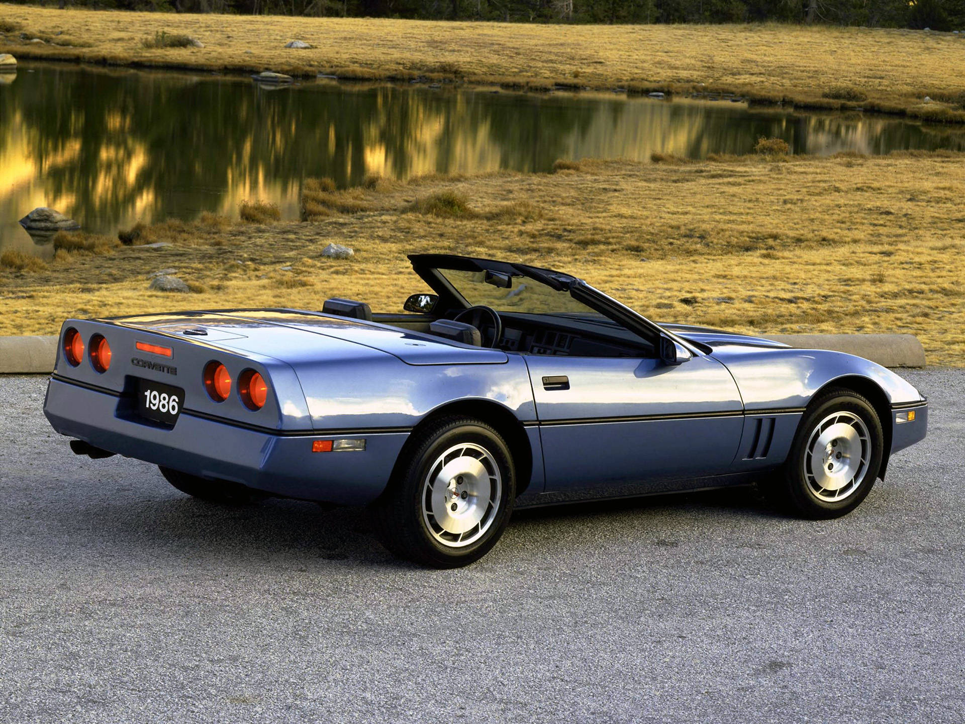 corvette 桌布 壁紙 — 一台時尚的藍色1986年雪佛蘭克爾維特敞篷車停在平靜的池塘旁，池塘倒映著金色的草和樹木。