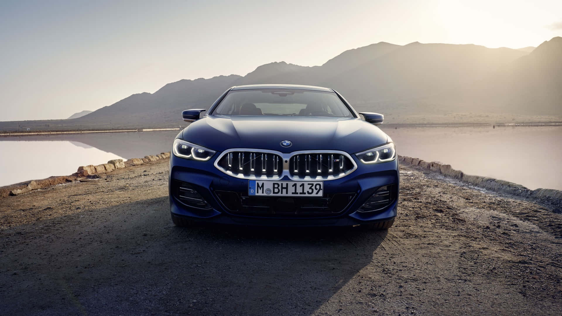 Bmw Cars 桌布 壁紙 — 一台時尚的藍色BMW 8系列雙門轎跑車停在碎石路上，旁邊是鹽灘，背景有山脈，夕陽西下。