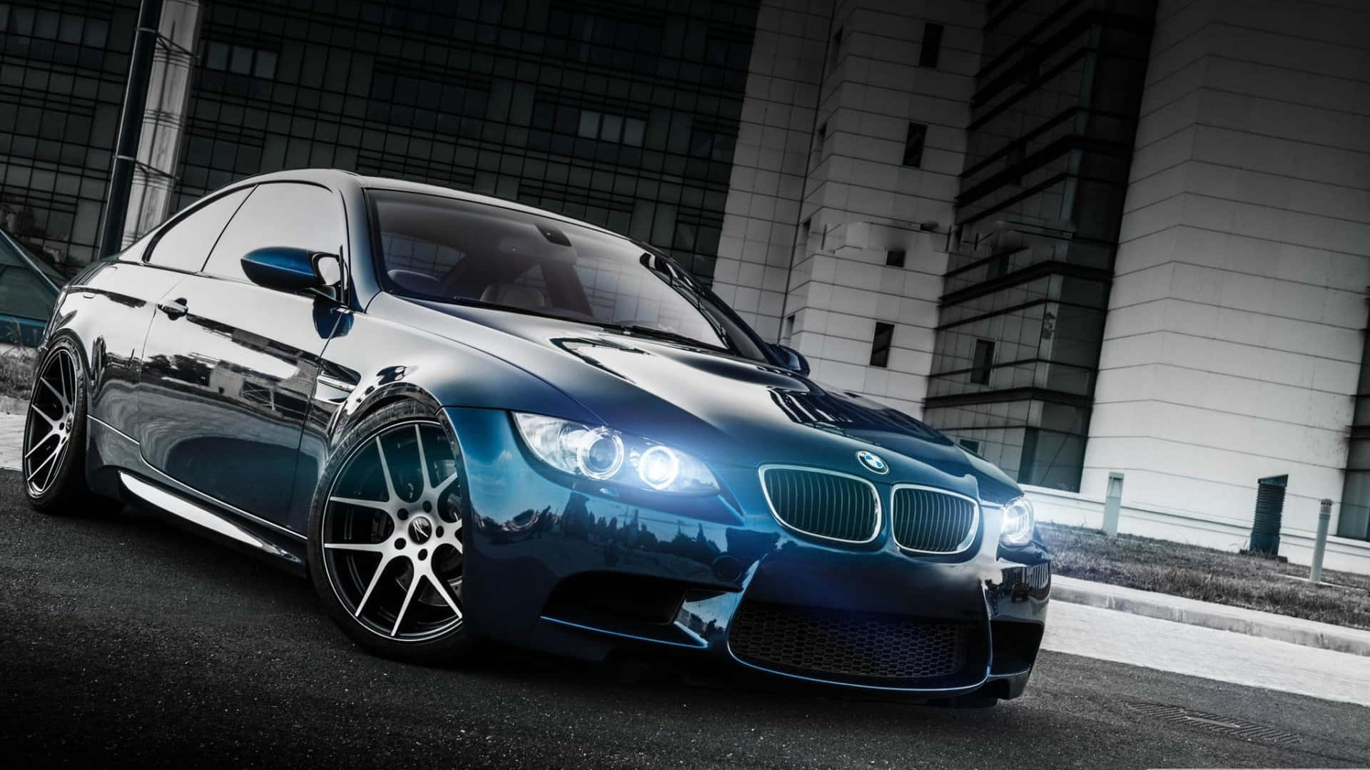1920x1080 Bmw 桌布 壁紙 — 一台時尚的藍色BMW M3雙門轎跑車在人造燈光下閃耀，背景是現代玻璃和混凝土建築構成的簡約都市景觀。