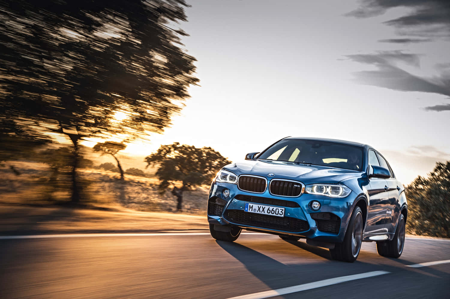 Bmw X6 M 桌布 壁紙 — 一台時尚的藍色BMW X6 M在日落時分沿著蜿蜒的道路疾馳，背景是模糊的樹木和金色的光線。
