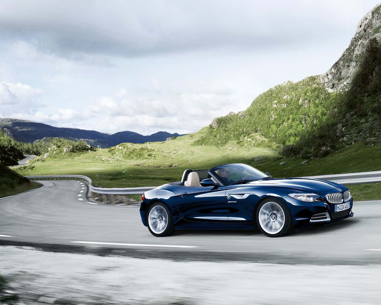 Bmw Z4 桌布 壁紙 — 一台時尚的藍色BMW Z4敞篷跑車在陰天之下，沿著蜿蜒的山路穿過翠綠的山丘飛馳。