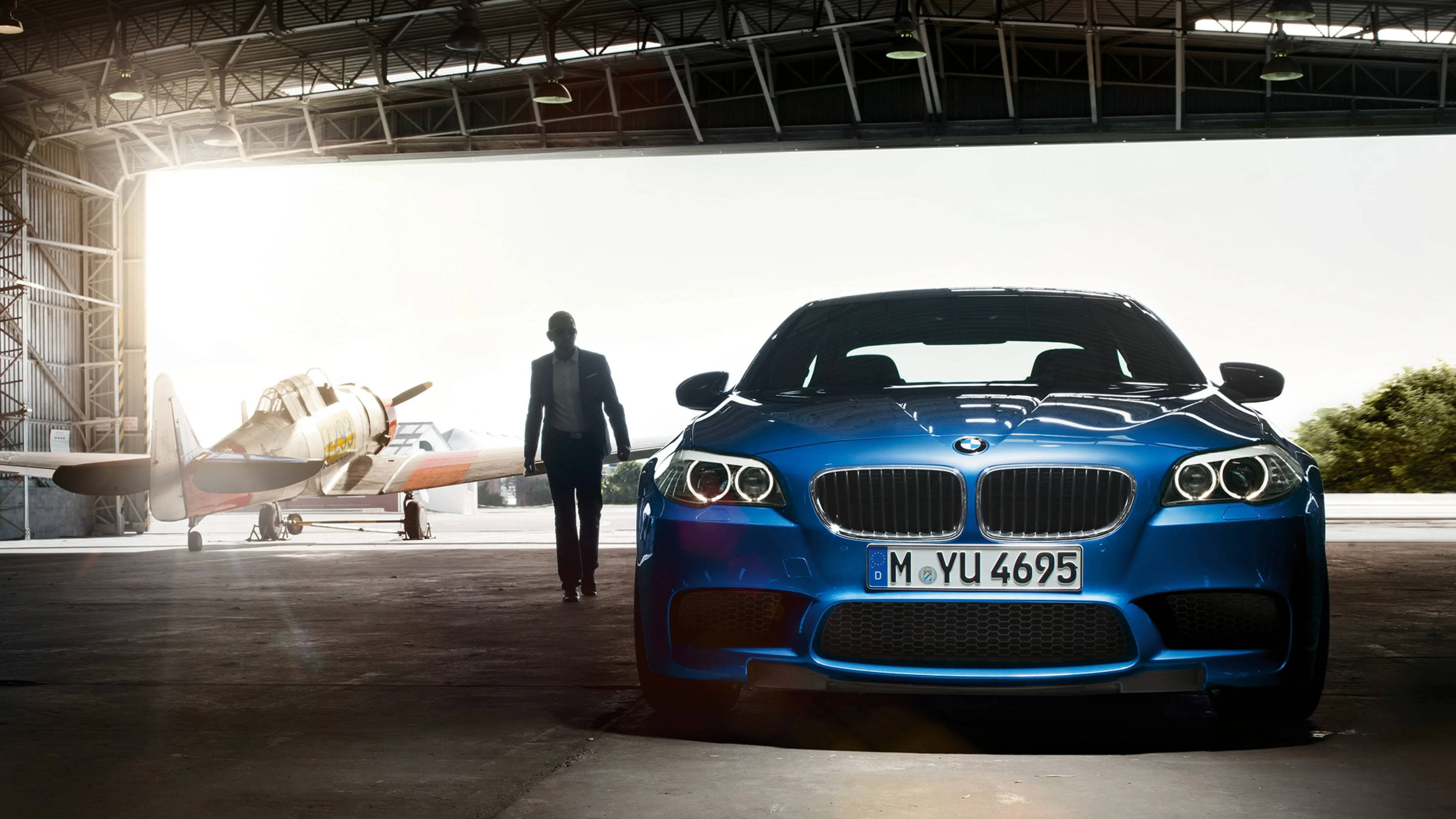 4K BMW 壁紙 桌布 壁紙 — 一台時尚的藍色BMW跑車在機庫內明亮燈光下閃閃發光，背景有一架復古飛機和一位身穿西裝的男子走向它。