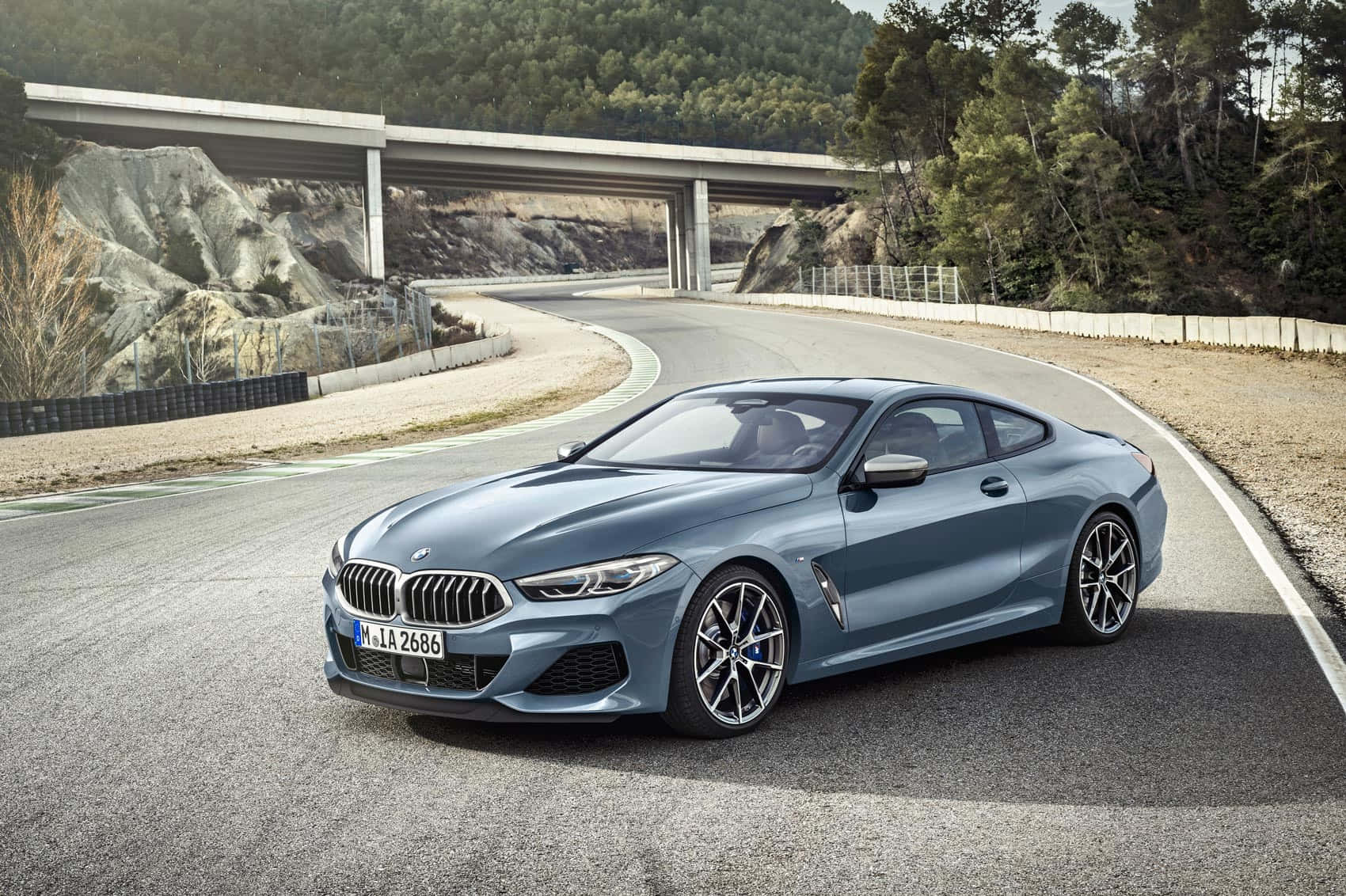 Bmw 8 Series 桌布 壁紙 — 一台時尚的藍色BMW轎跑車在混凝土高架橋下蜿蜒的山路上行駛，兩旁是森林覆蓋的山丘。
