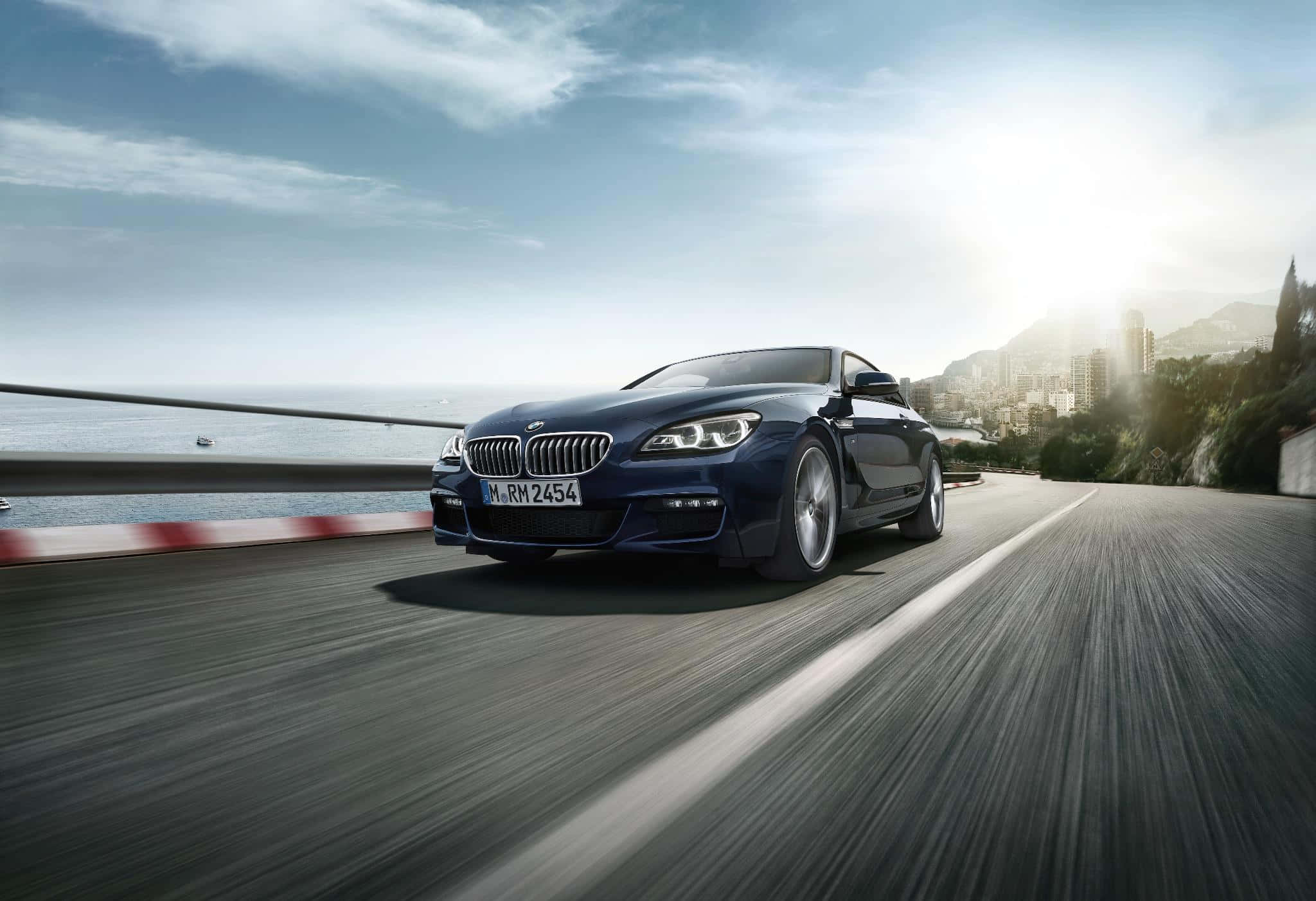 BMW 動圖 桌布 壁紙 — 一台時尚的藍色BMW雙門跑車沿著海岸公路疾馳，背景是海洋和陽光普照的懸崖，帶有動態模糊效果。