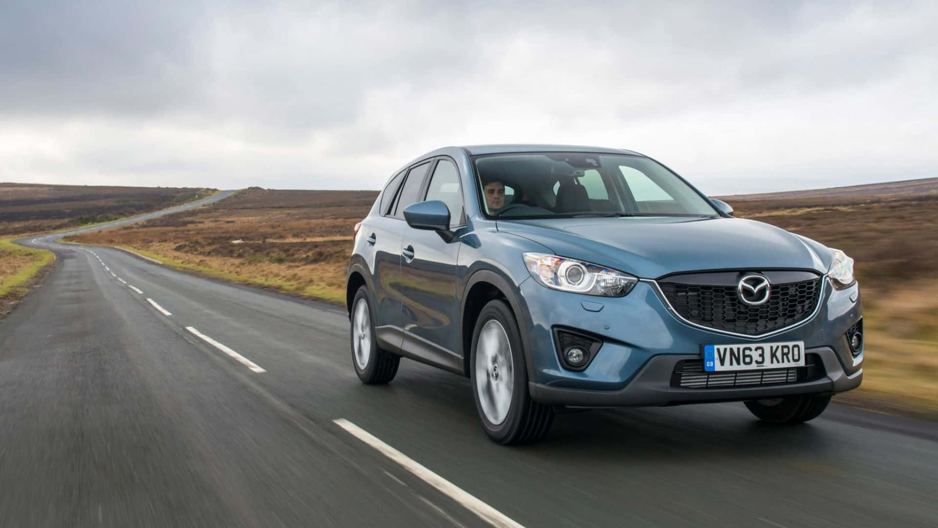 Mazda Cx-5 桌布 壁紙 — 一台時尚的藍色Mazda CX-5在陰天下行駛於蜿蜒的鄉村道路，穿過開闊的荒野。