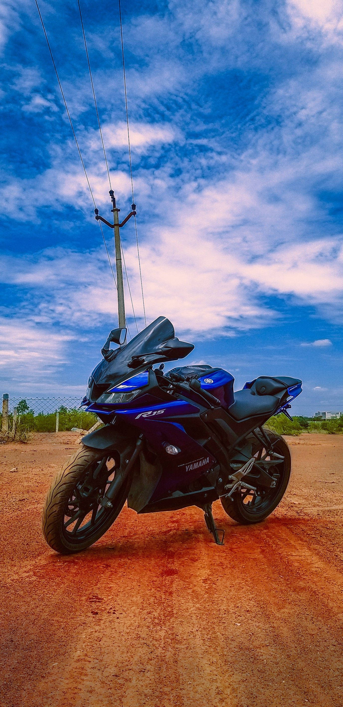桌布 壁紙 — 一台時尚的藍色Yamaha R15機車停在塵土飛揚的紅色泥土路上，頭頂是湛藍的天空，點綴著縷縷白雲和電線。