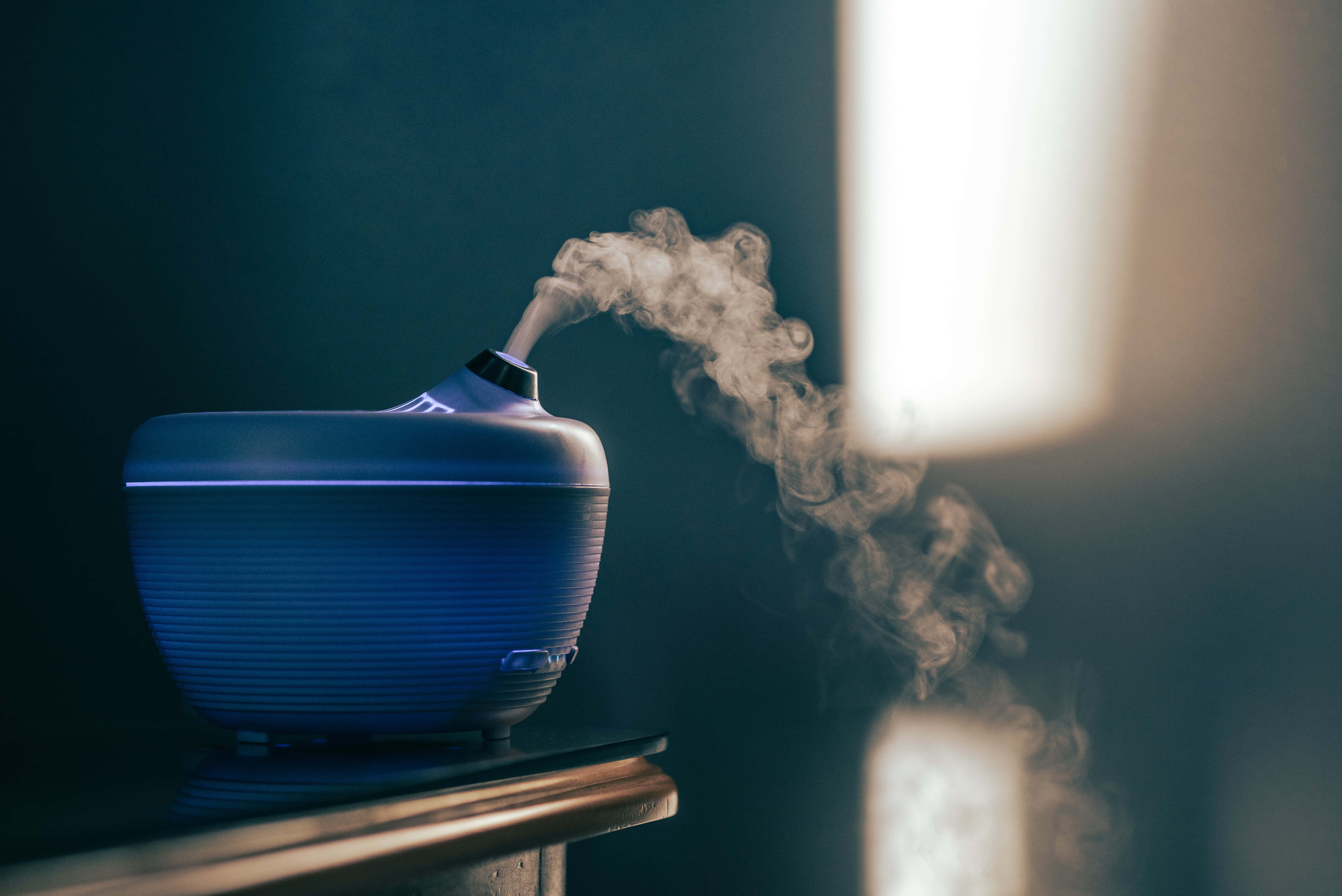 Smokey 桌布 壁紙 — 一台時尚的藍色加濕器在昏暗的房間中釋放出溫和的霧氣，柔和自然光透過窗戶照射進來。