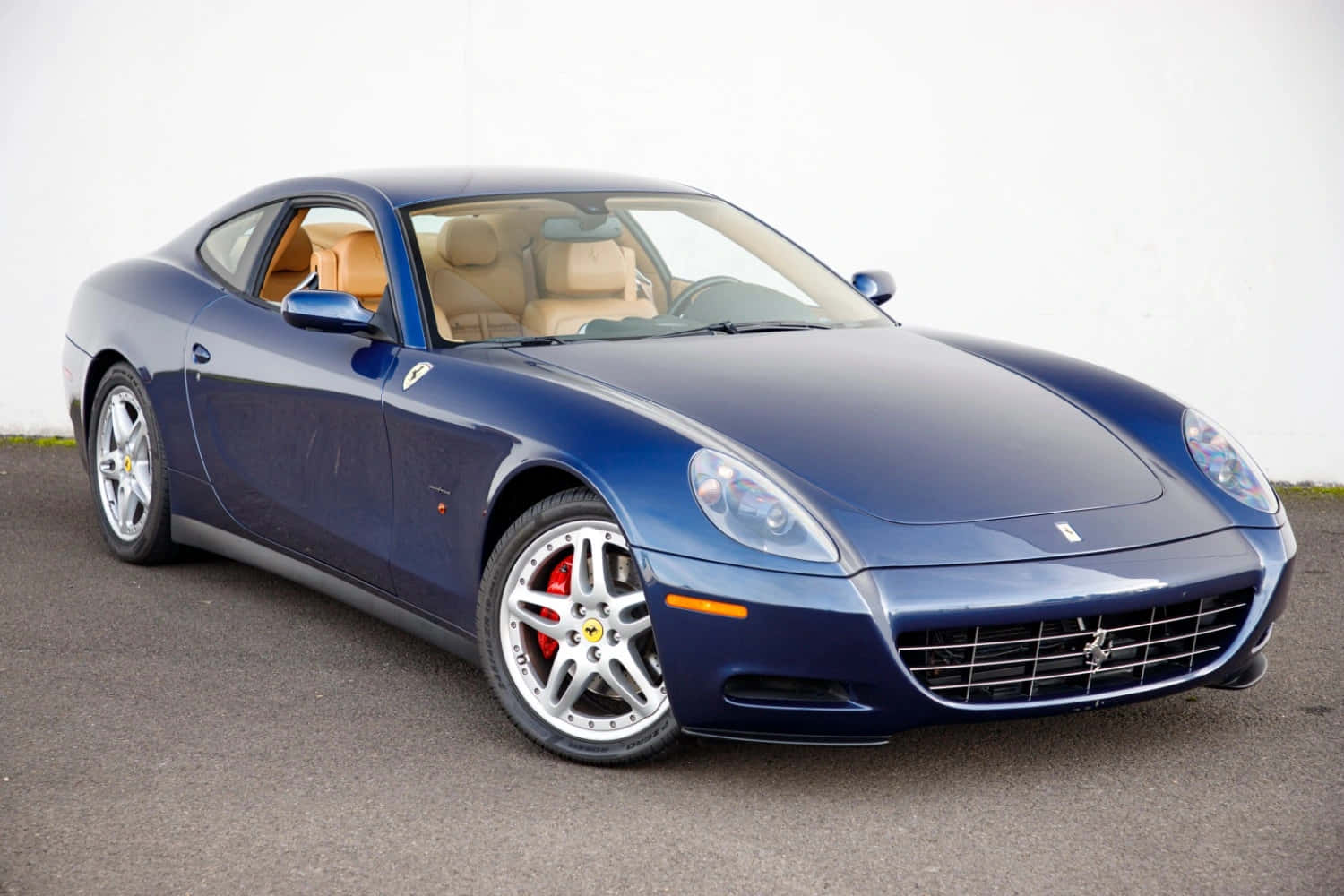 Ferrari 612 Scaglietti 桌布 壁紙 — 一台時尚的藍色法拉利雙門跑車，配備淺褐色皮革內裝，停放在柏油路面上，背景是純白色牆壁。