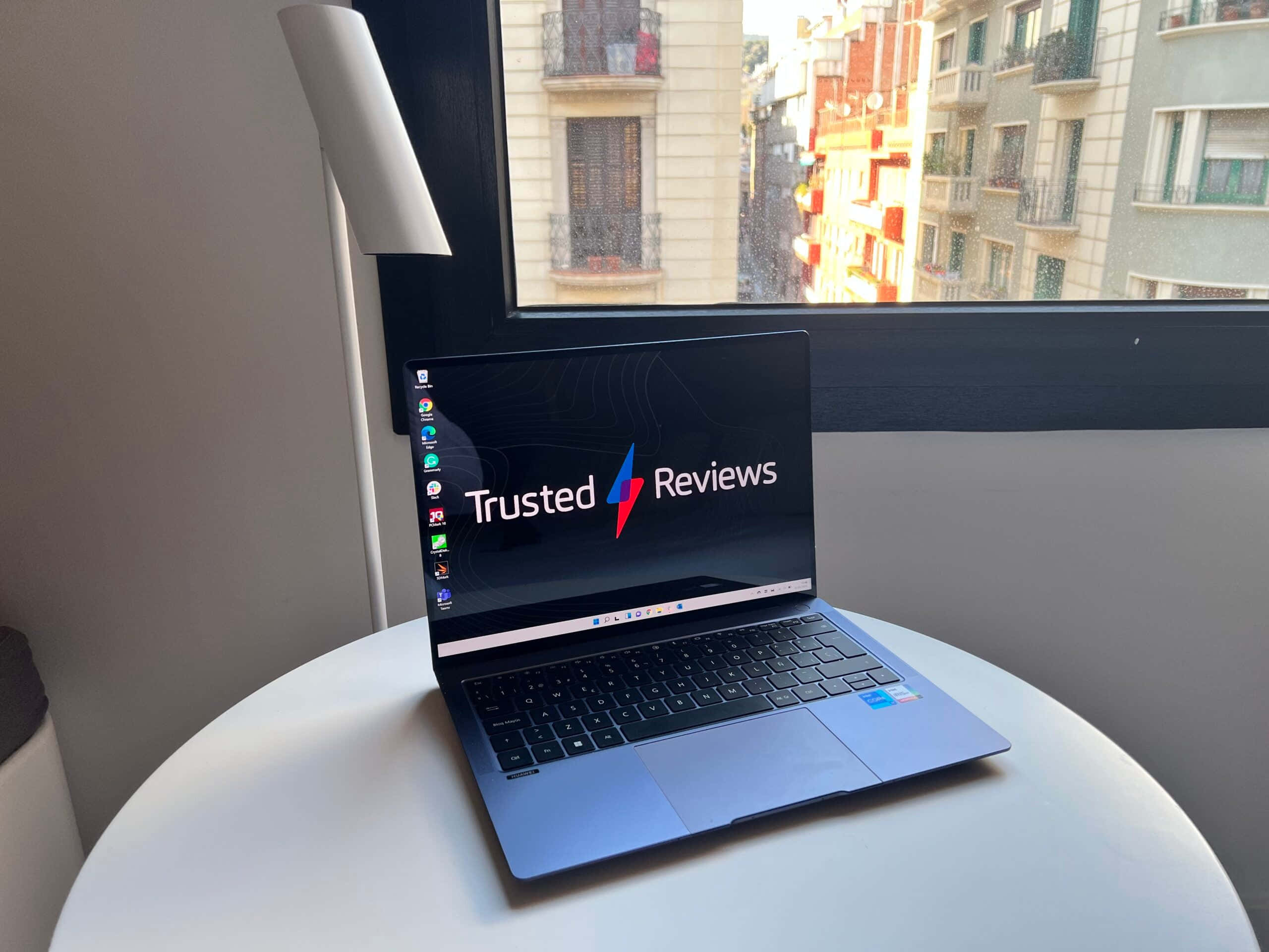 筆電 桌布 壁紙 — 一台時尚的藍色筆記型電腦顯示著 Trusted Reviews，放在靠近窗戶的白色圓桌上，窗外可俯瞰城市建築。