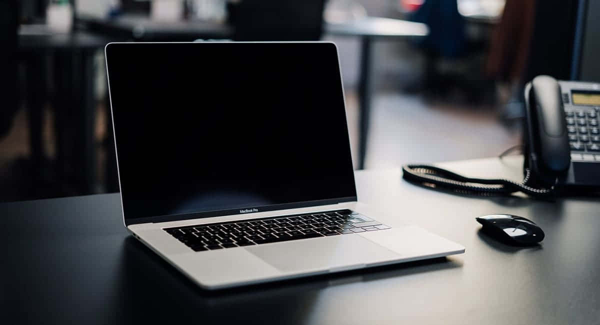 Black Macbook 桌布 壁紙 — 一台時尚的銀色 MacBook Pro 打開放在深色桌子上，旁邊有一個黑色滑鼠和一個座機電話，背景是柔和模糊的辦公室環境。