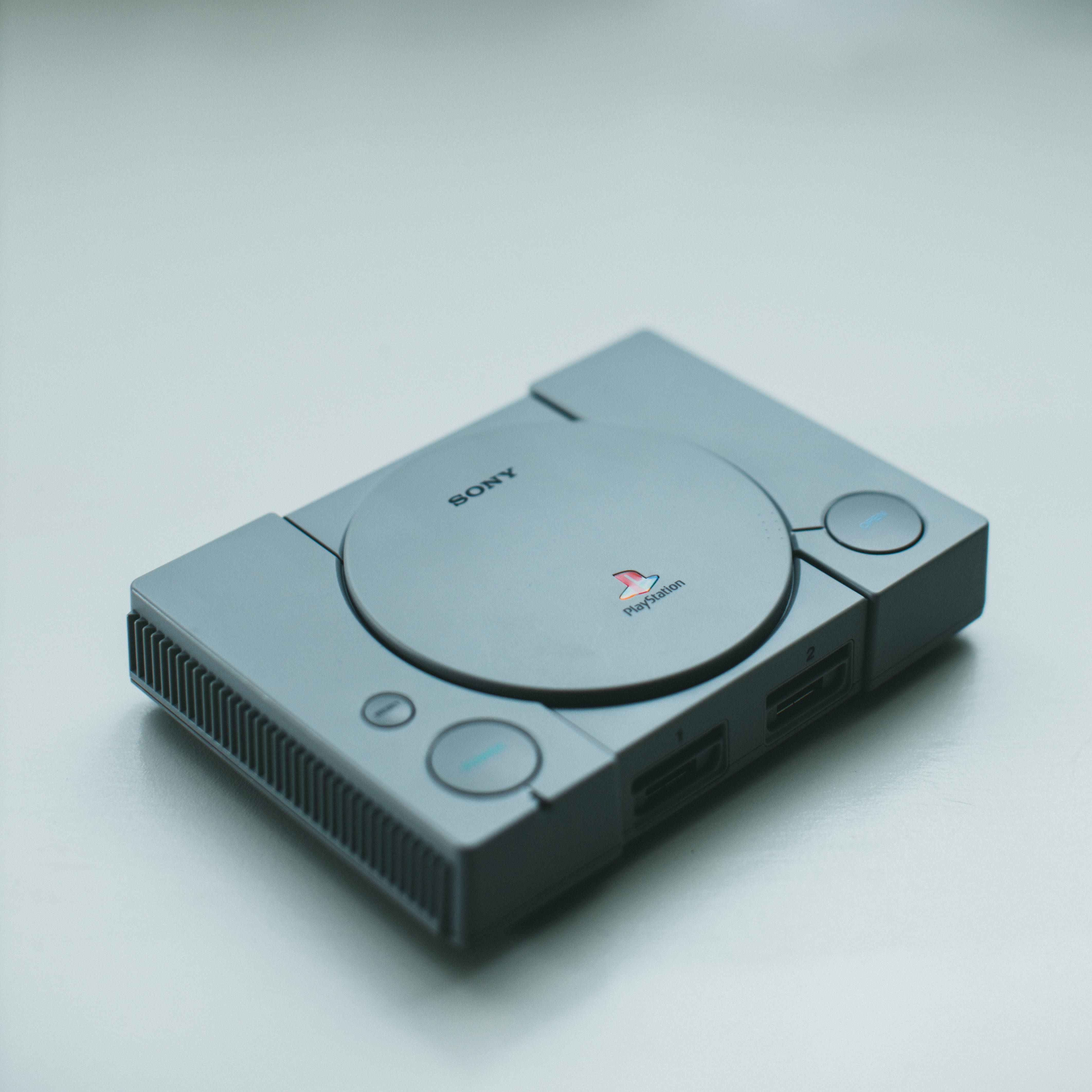 Sony 桌布 壁紙 — 一台時尚的銀色 PlayStation 1 主機放置在簡約的白色表面上，柔和的光線突顯其標誌性的圓形設計。