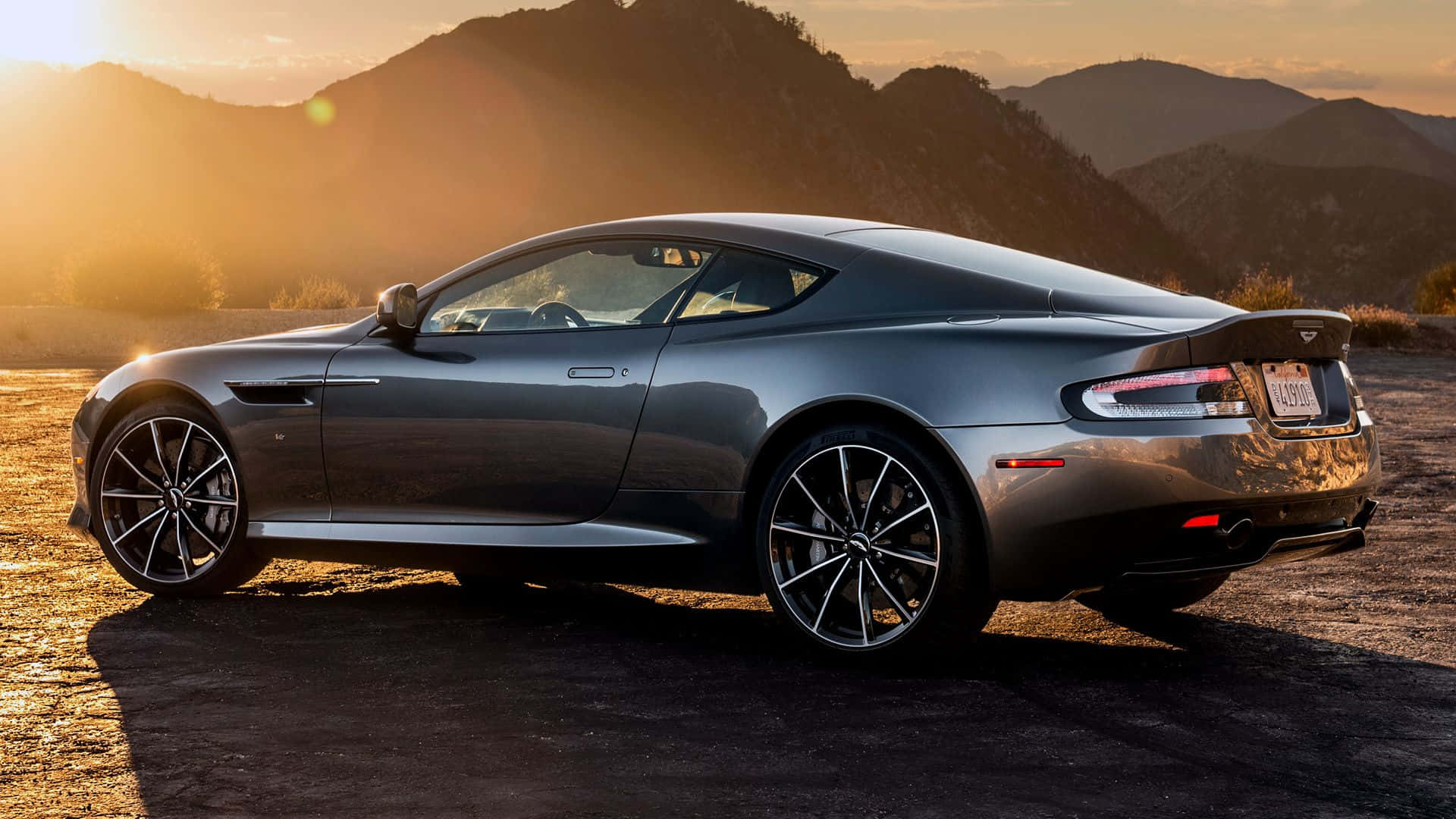 Aston Martin Db9 桌布 壁紙 — 一台時尚的銀色Aston Martin跑車停泊在塵土飛揚的道路上，群山沐浴在金色的日落光芒中。