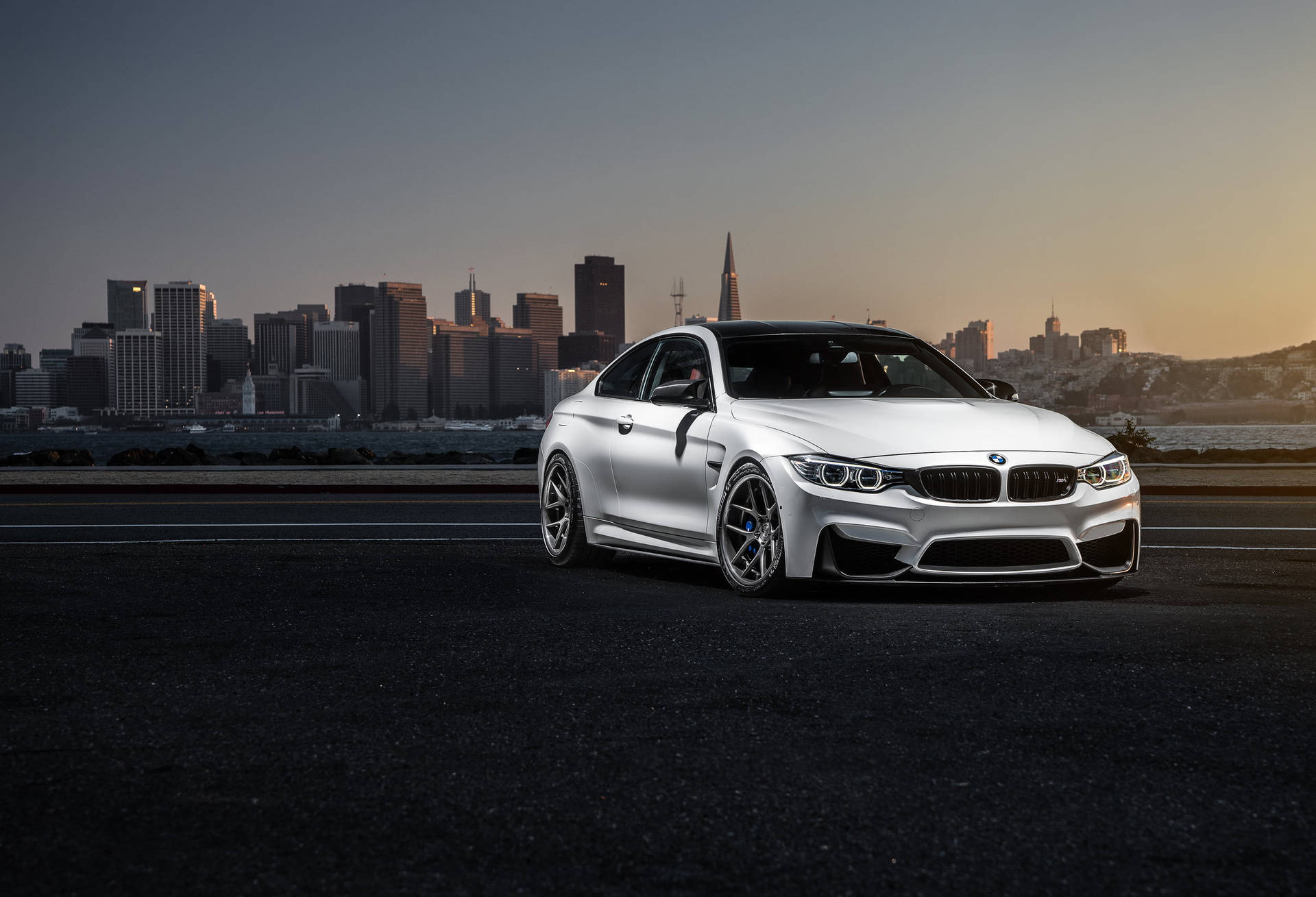 銀色BMW M4雙門跑車停泊在道路上，夕陽下的城市天際線。 — BMW 動圖 壁紙